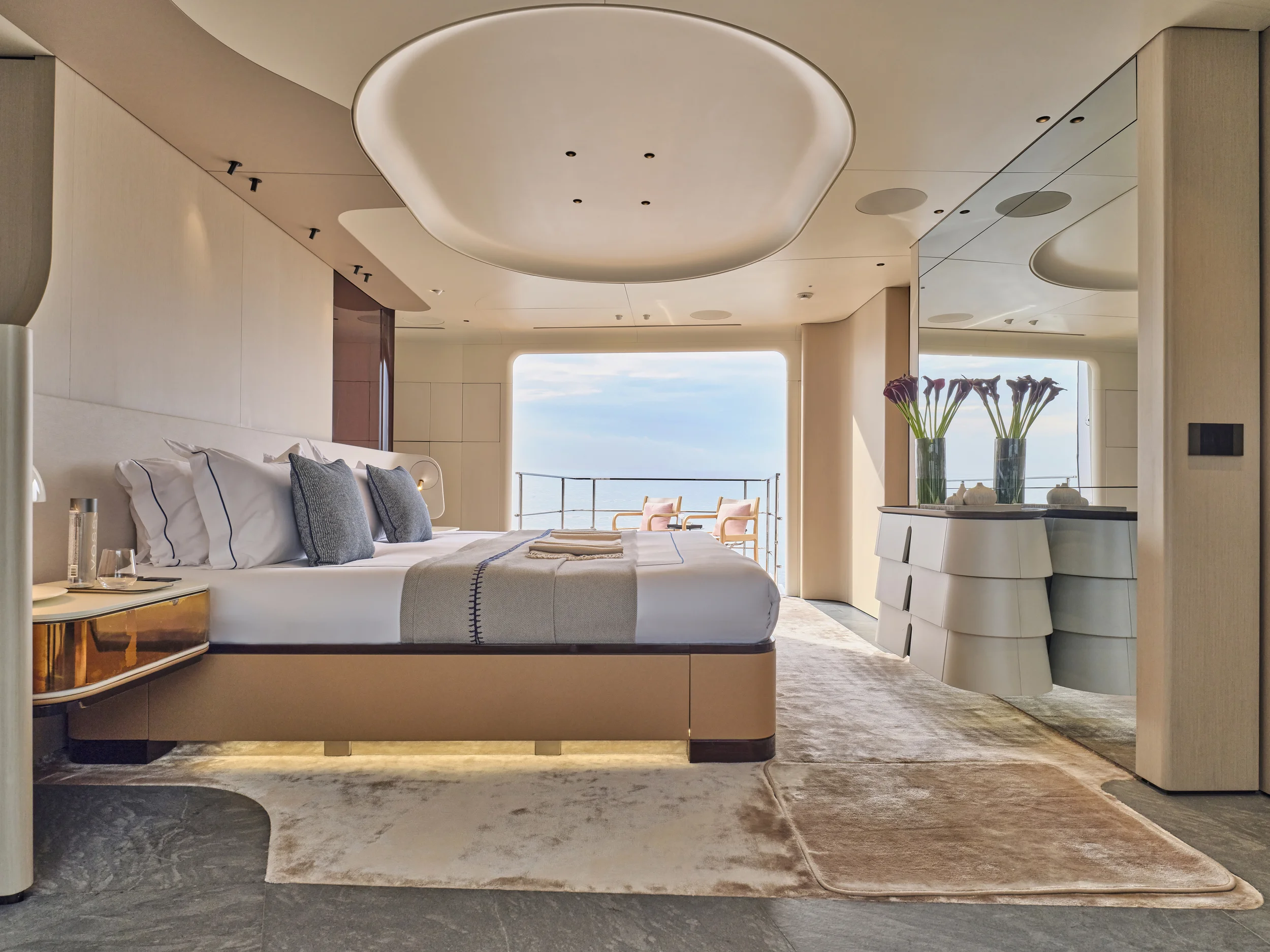 Luxury hotel room with a large bed, pillows, and a balcony with chairs overlooking the ocean, decorated with vases of flowers and modern furniture.