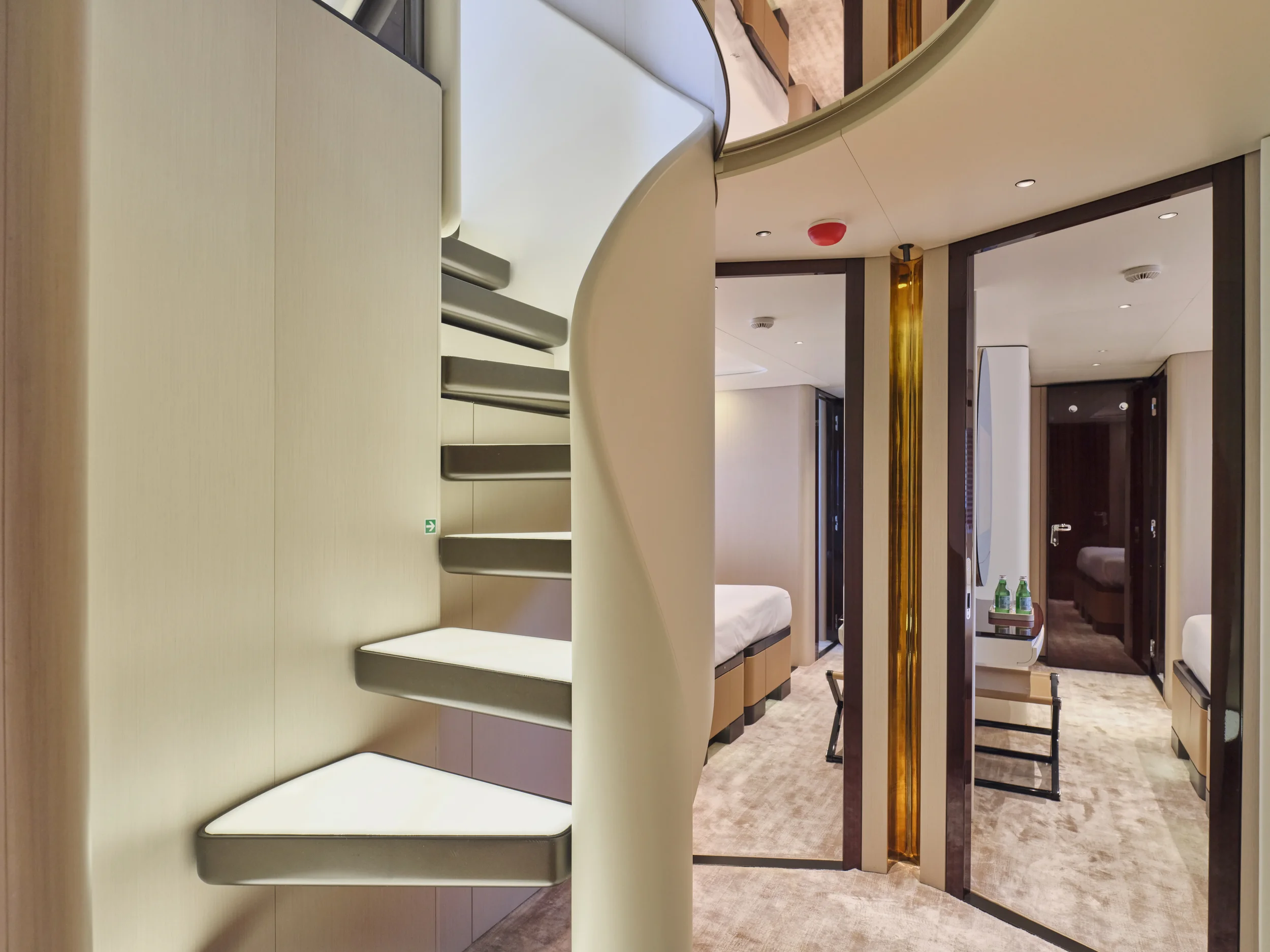 View of an interior hotel suite with a staircase to a lofted area on the left and a sitting and sleeping area on the right, featuring beds, chairs, and a tray with bottles of water.