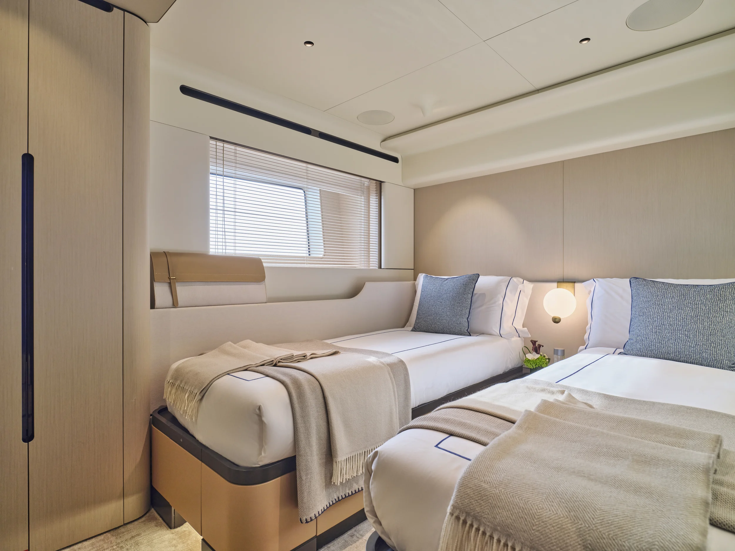 Modern hotel room with two beds dressed in white linens, beige and blue pillows, beige throws, light wood furniture, a window with blinds, and a wall-mounted lamp.
