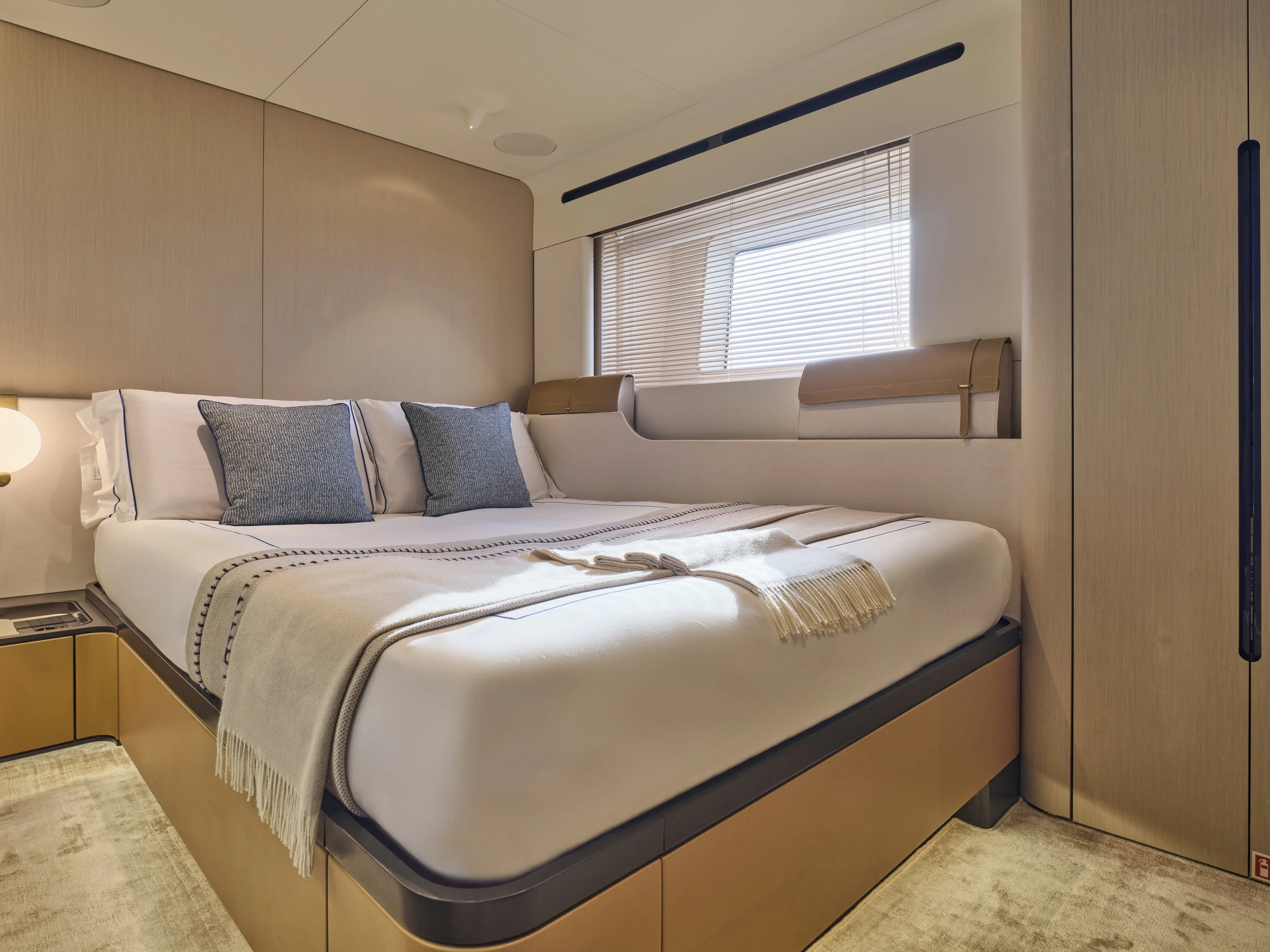 Modern bedroom with a large bed, gray pillows, beige throw blanket, window with blinds, beige walls, and minimal decor.