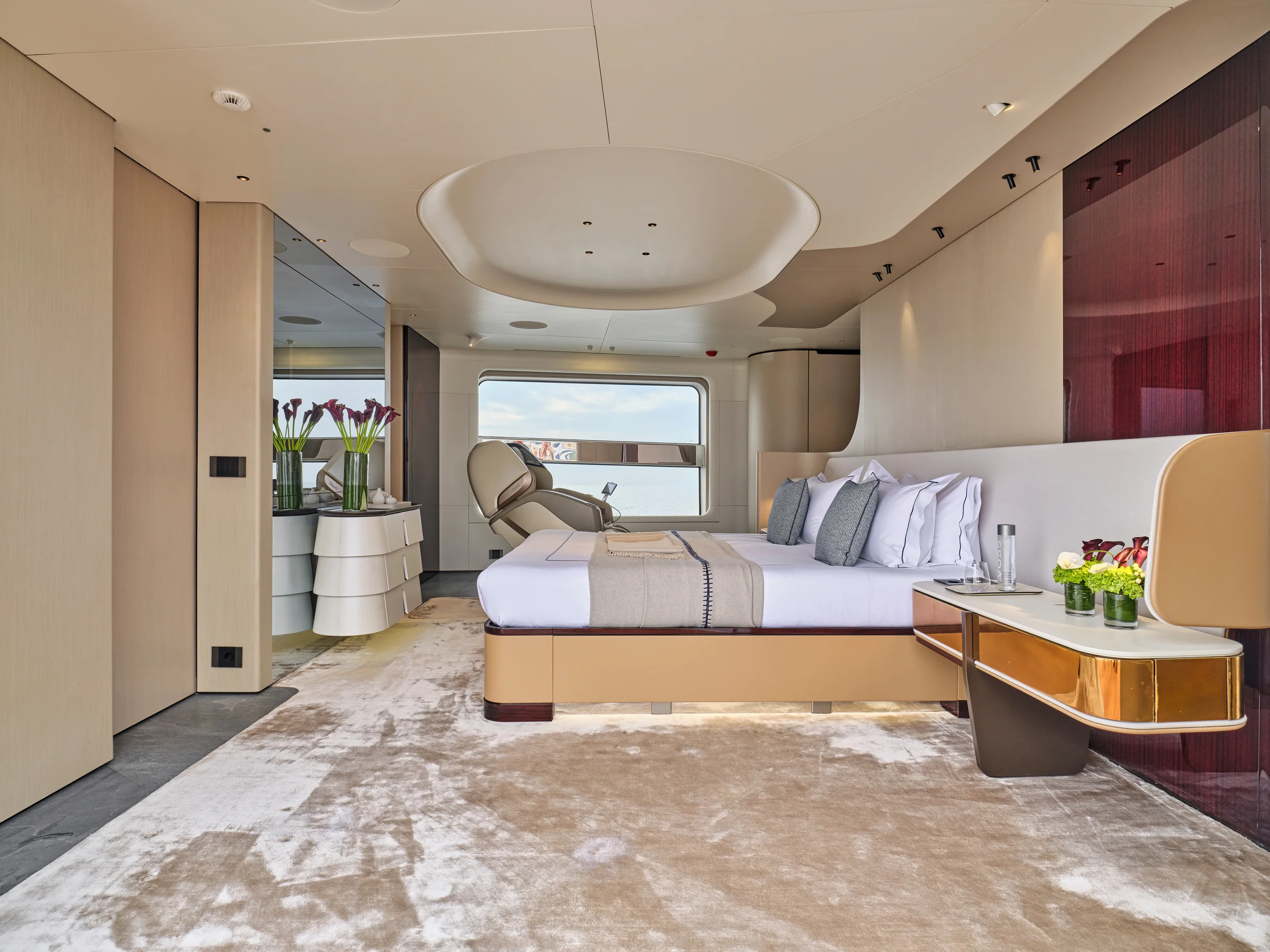 Luxurious hotel bedroom with a large bed, side tables with flowers, a window overlooking the water, and a reclining chair.