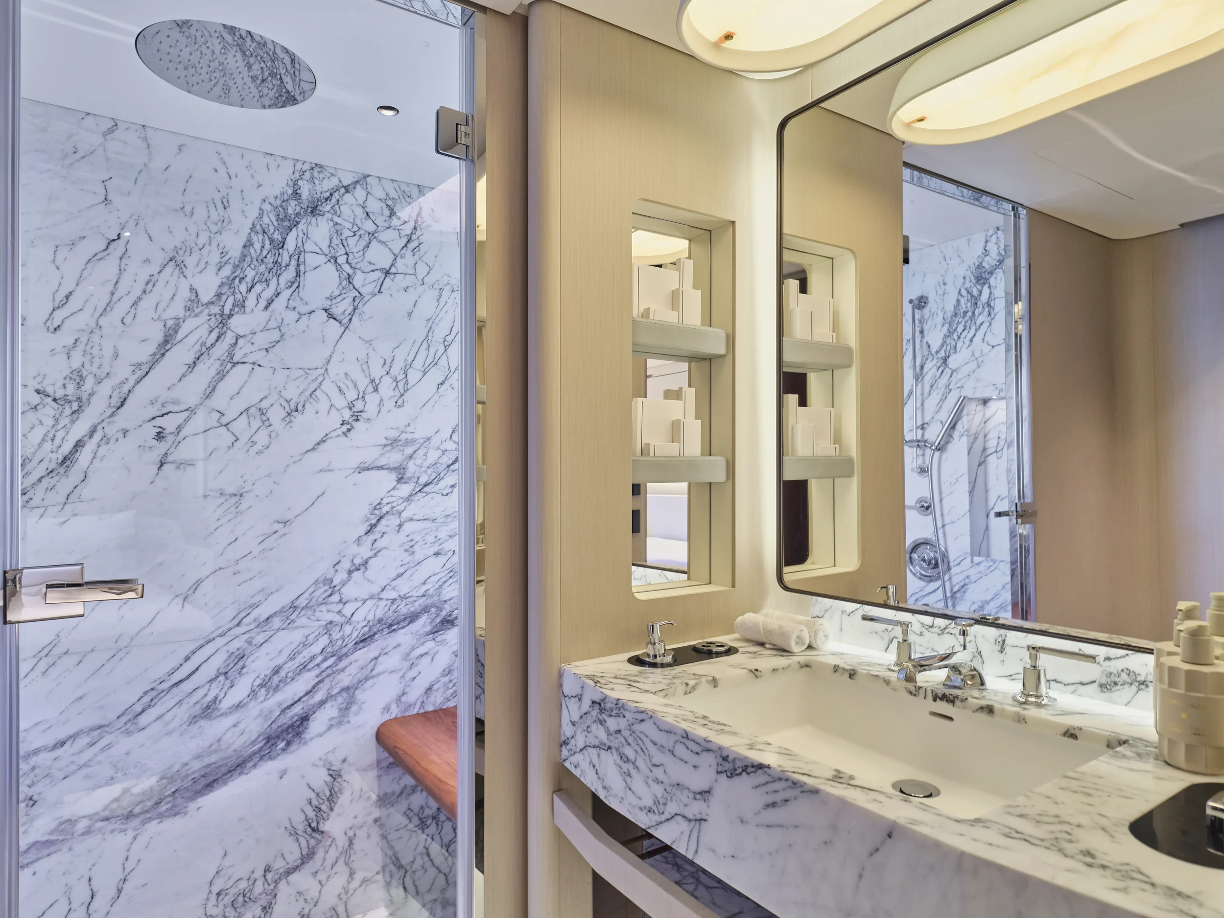 A luxurious bathroom with a marble countertop and backsplash, a large mirror, a wooden shelf with toiletries, and a walk-in shower with marble walls, a rainfall showerhead, and a wooden bench.