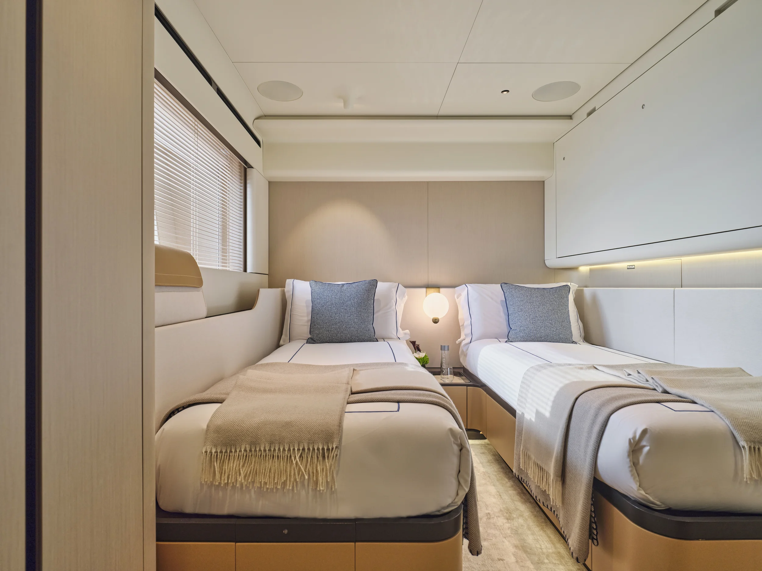 A modern bedroom with two twin beds, beige throws, gray pillows, a nightstand with a lamp, water bottle, and flowers, a window with blinds, and neutral-colored walls.