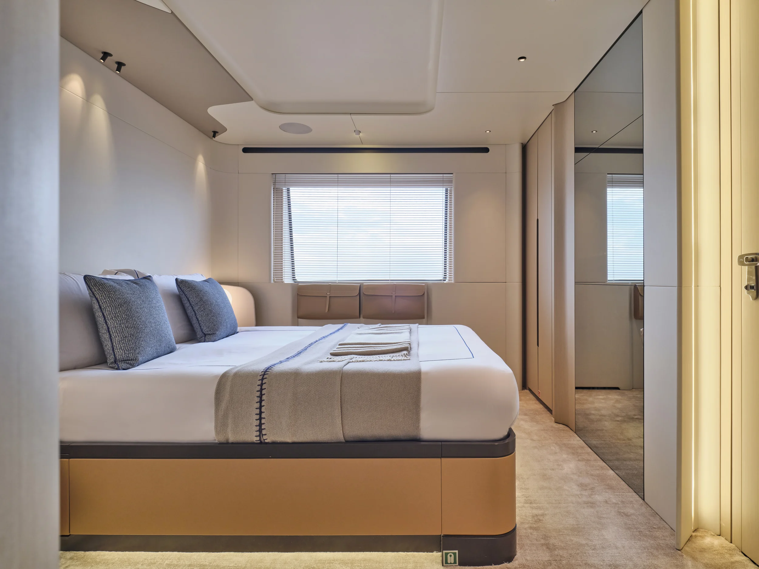 Modern hotel room with a large bed, gray pillows, beige walls, a window with blinds, and mirrored closet doors.