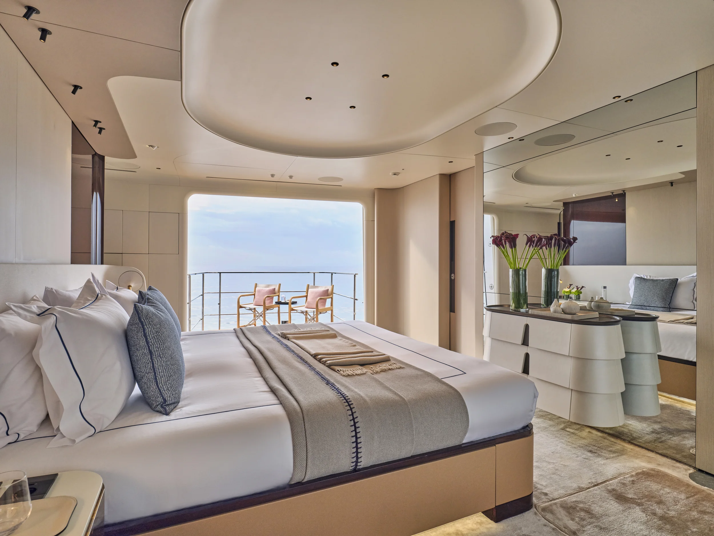 Luxury hotel room with a large bed, decorative pillows, a blanket, a bedside table with a glass of water, and a balcony with two chairs overlooking the sky and ocean.