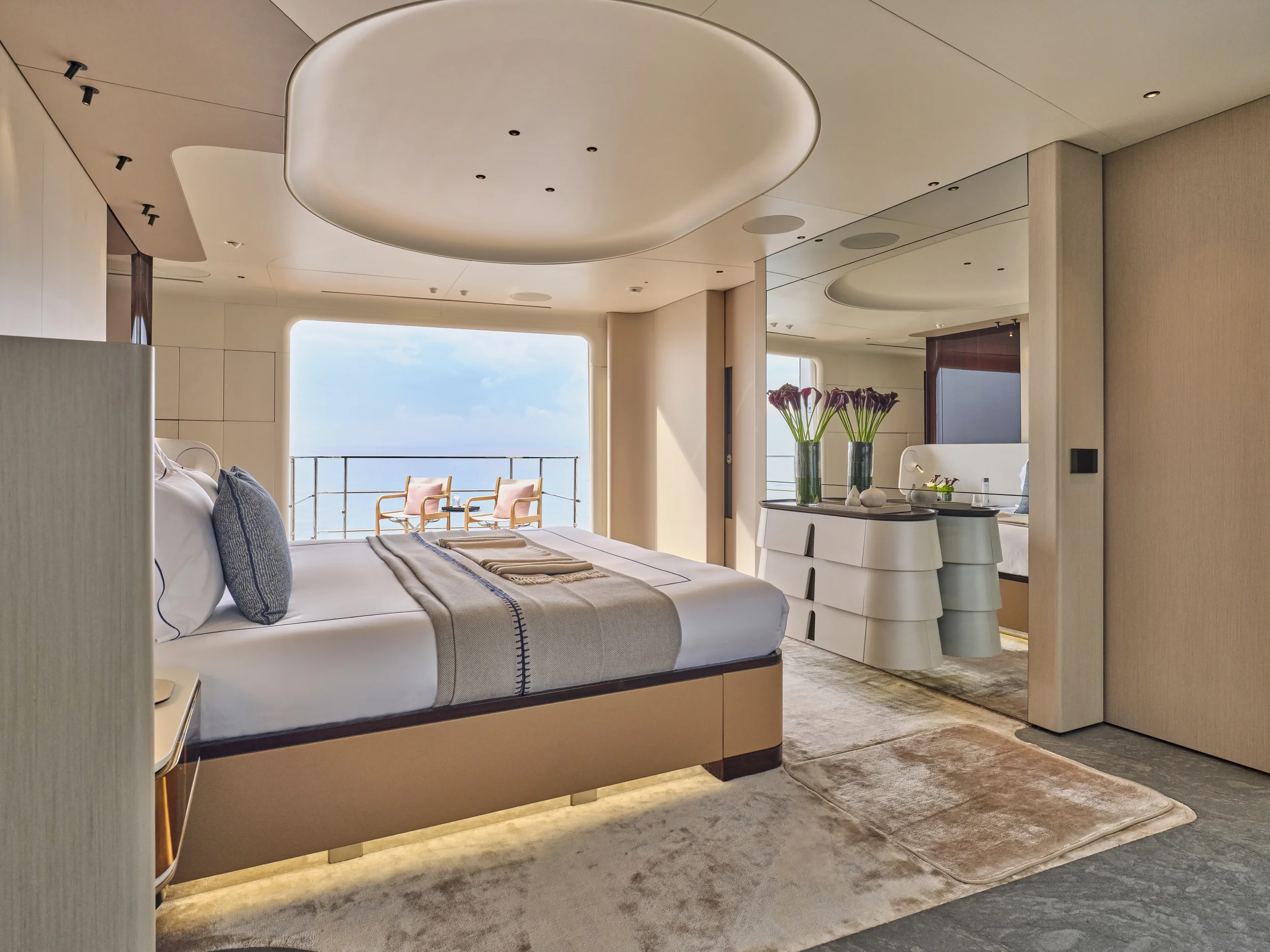 Luxurious hotel bedroom with a large bed, sitting area outside on the balcony with two wooden chairs and cushions, and contemporary decor including vases with flowers and a dresser.