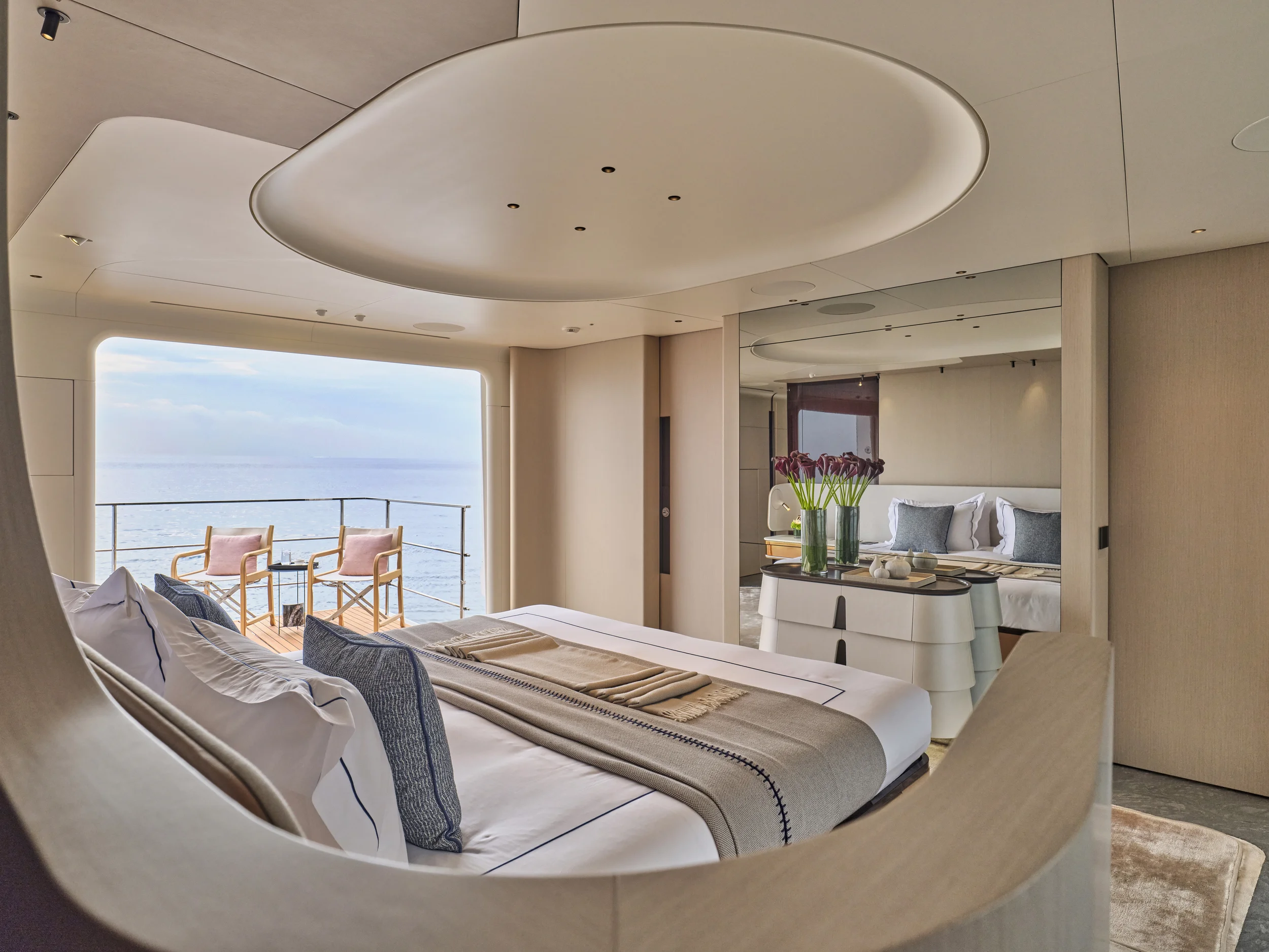 Luxury bedroom with a large bed, decorative pillows, and a beige bed runner, overlooking a balcony with two chairs and the ocean in the background.