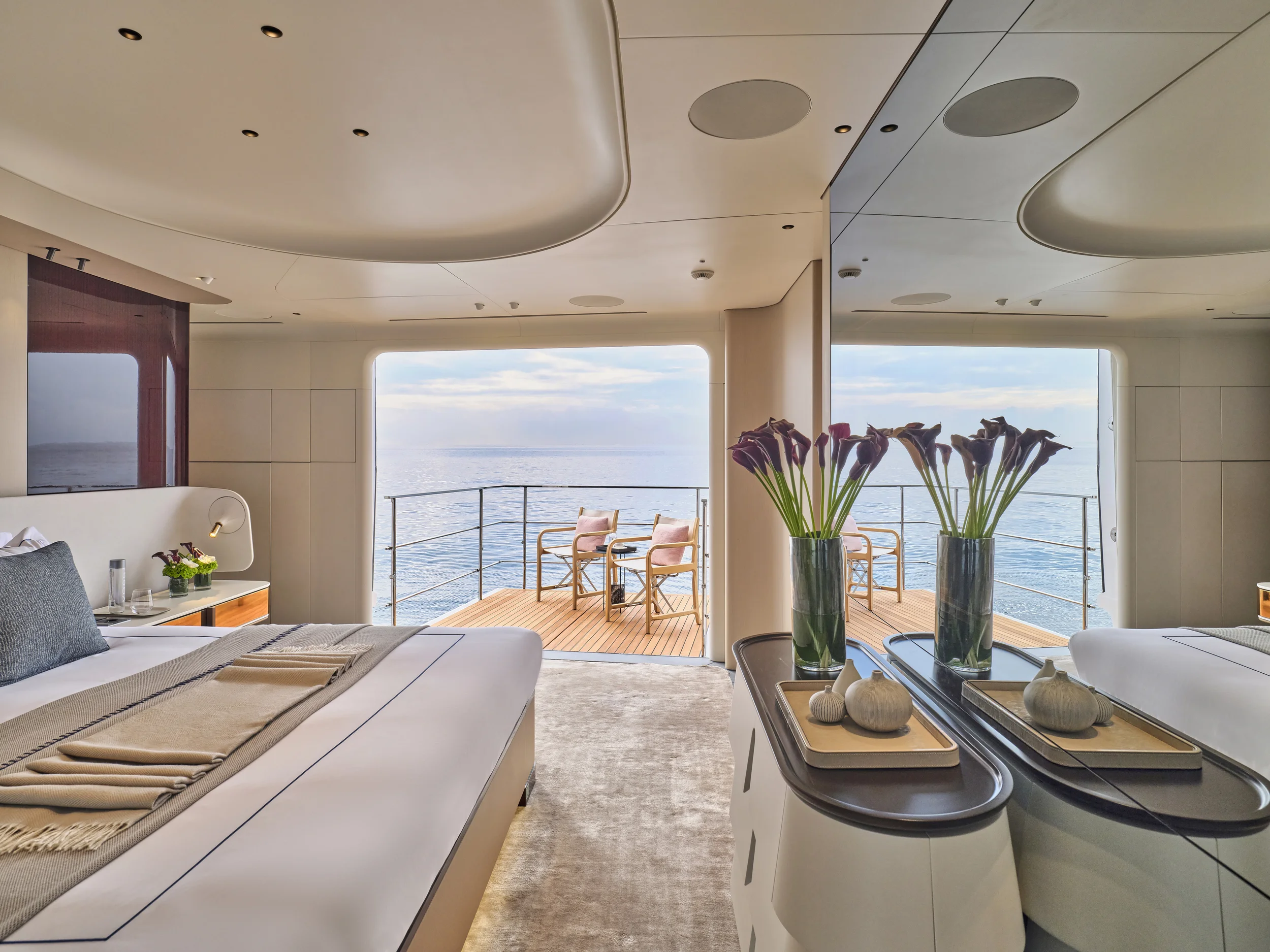 Luxury cruise ship bedroom with a balcony view of the ocean, decorated with vases of flowers, a bed with linen, and a small sitting area outside on the balcony.