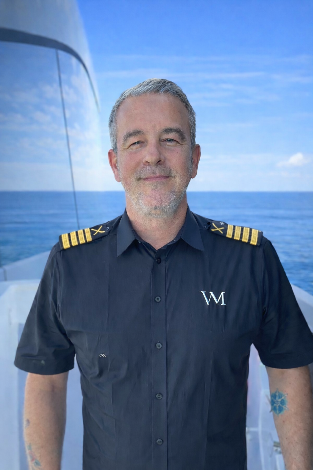 A smiling man in a pilot uniform with shoulder epaulets, standing outdoors near water with a boat in the background.