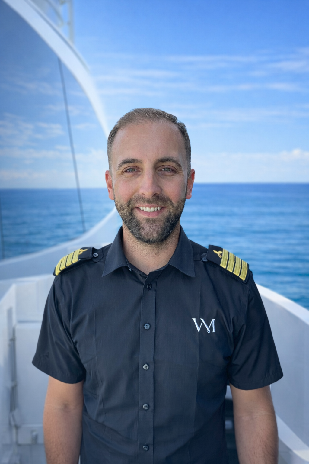 Man in a navy uniform with captain's epaulets standing in front of a boat and water