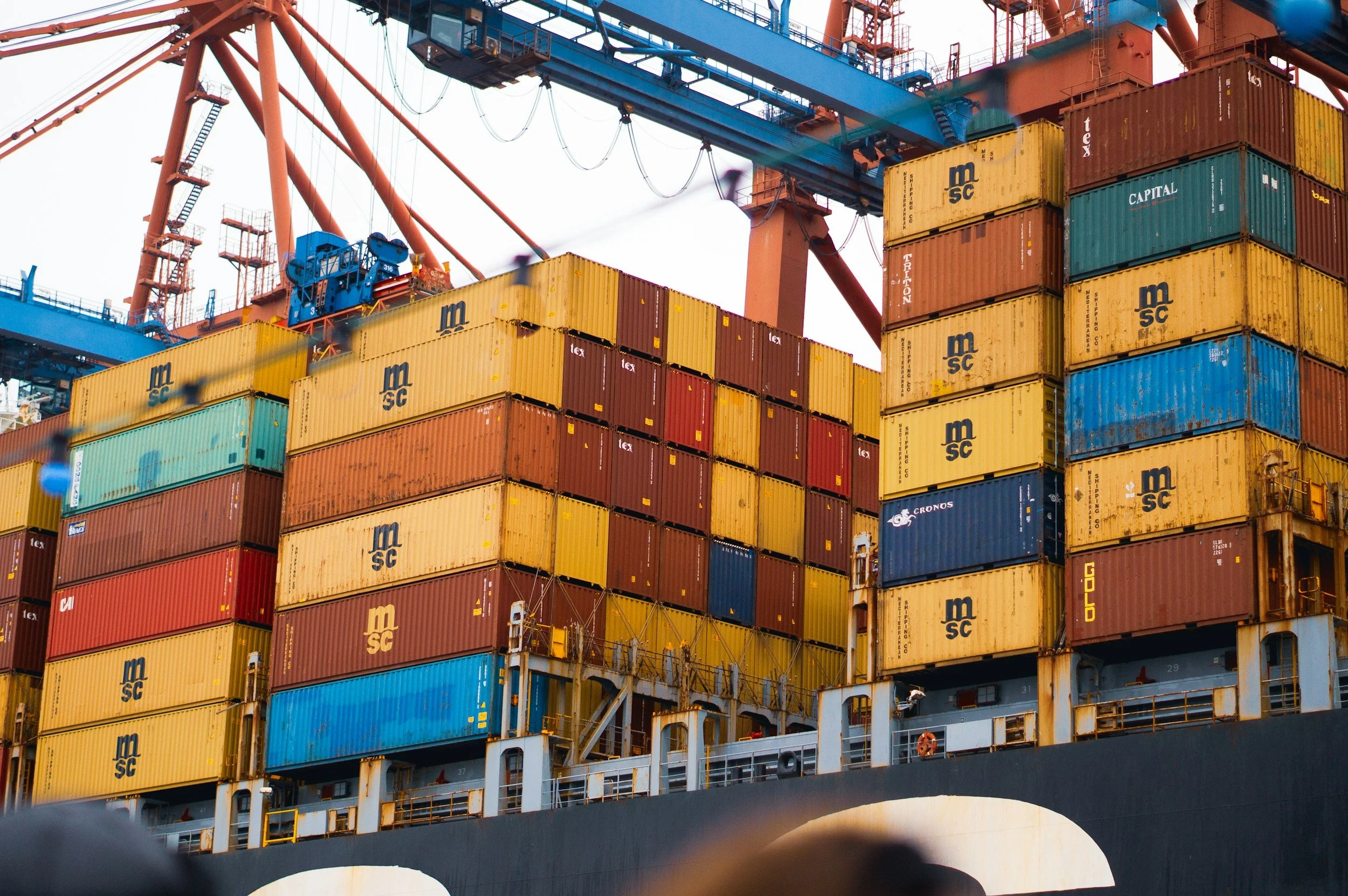 A large container ship loaded with multicolored shipping containers, with a focus on yellow, blue, red, and green containers bearing various company logos, docked at a port.