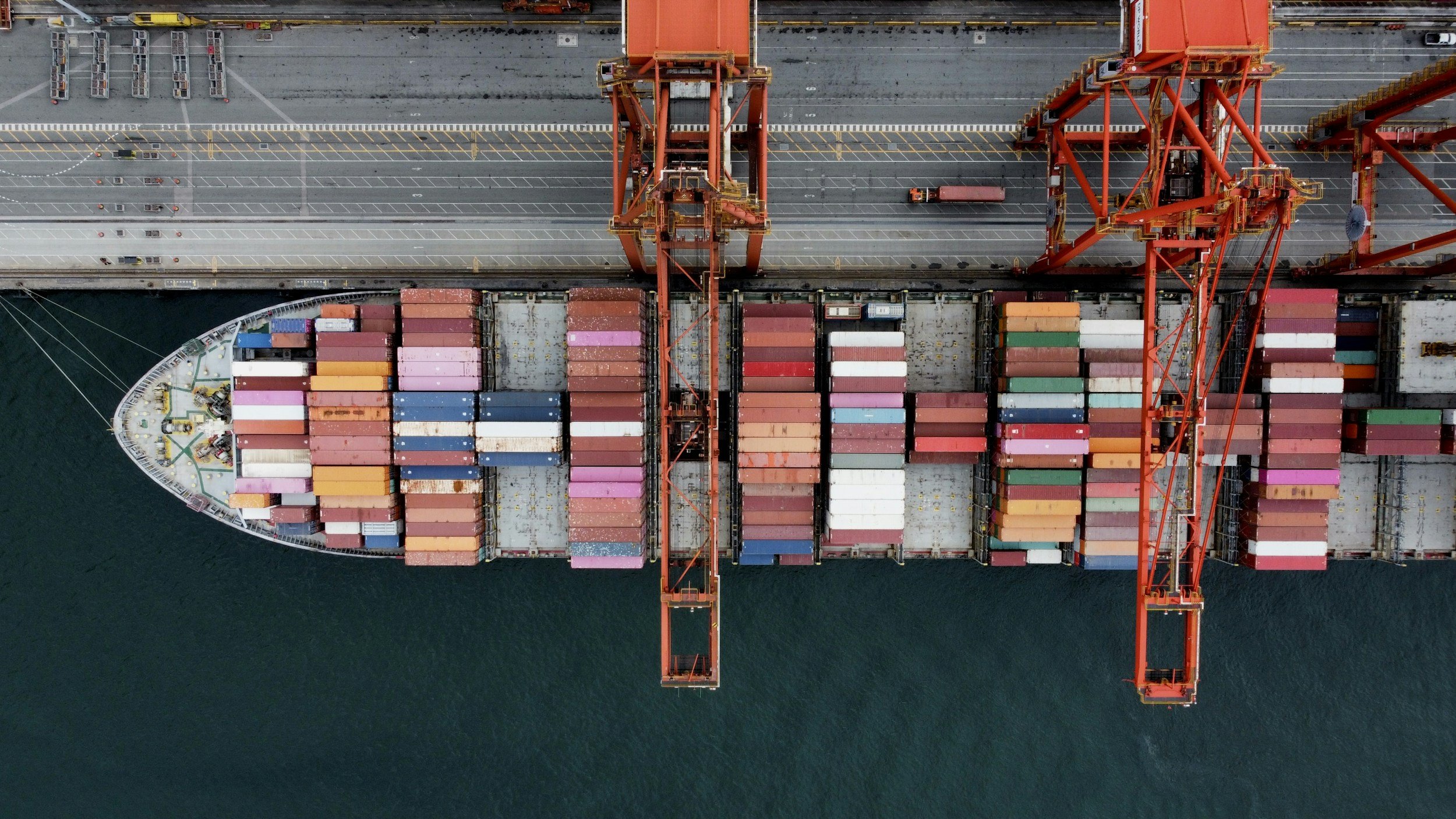 Aerial view of a cargo ship loaded with multicolored containers, docked at a port with cranes.