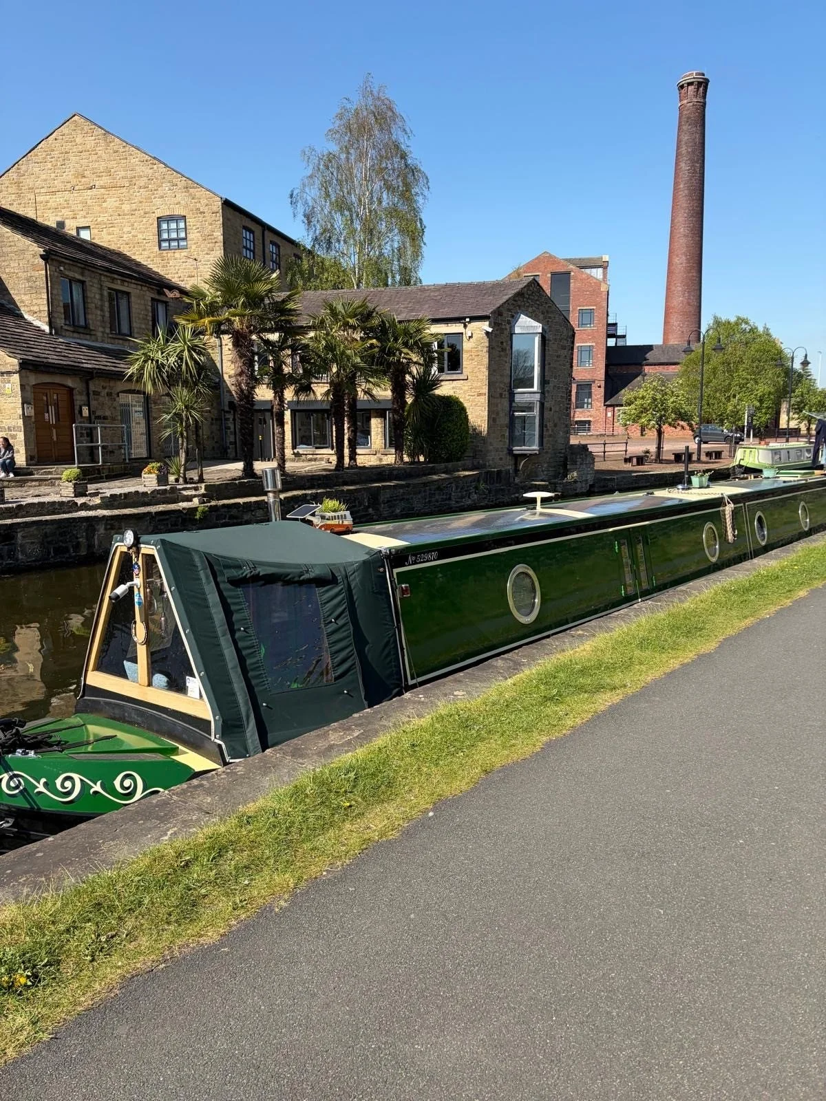 A narrow green canal boat moored alongside a sidewalk next to a canal, with historic buildings, trees, and a clear blue sky in the background.