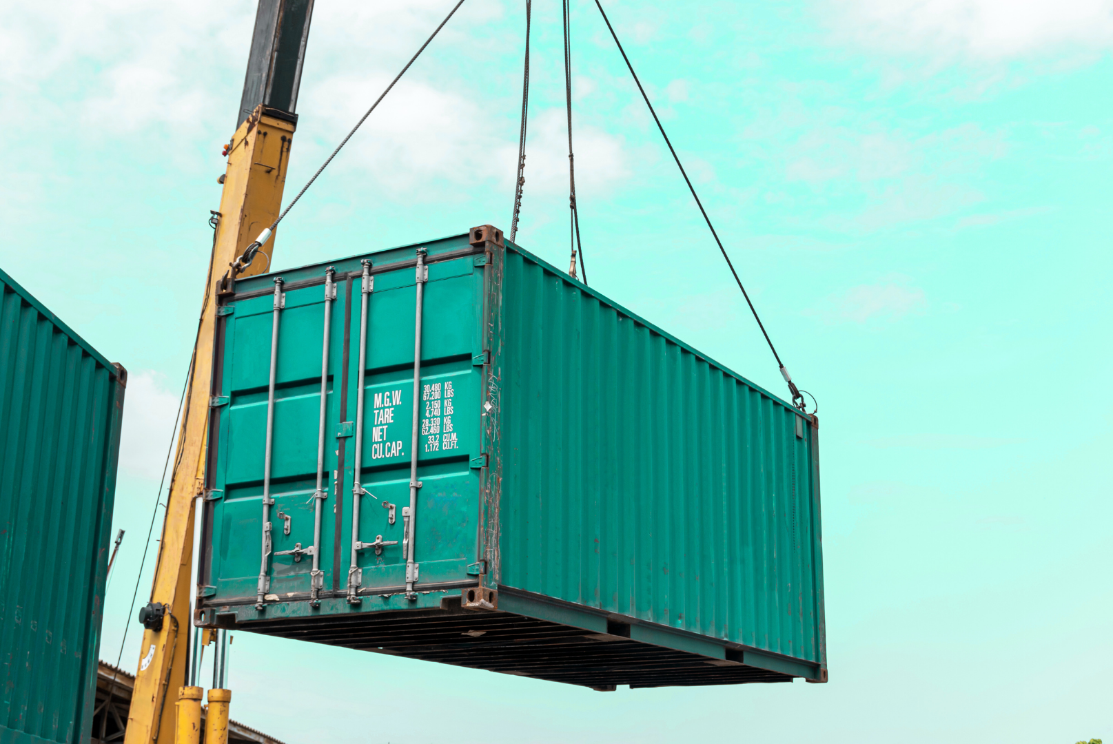 A large green cargo container being hoisted by a crane against a partly cloudy sky.