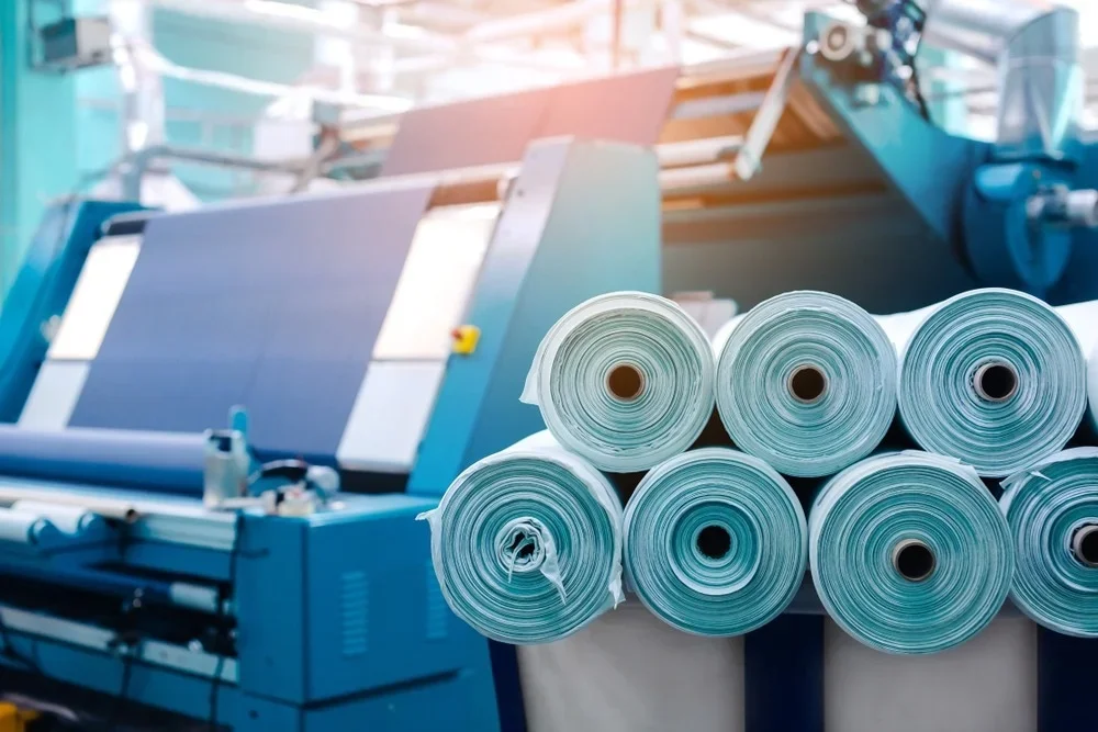 Multiple rolls of blue and white printing paper stacked in front of a large industrial printing machine.
