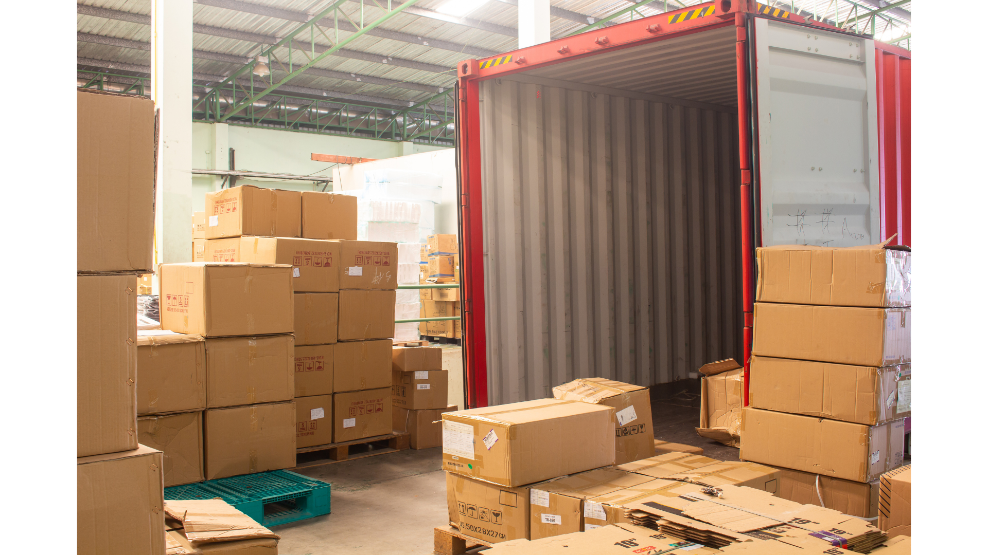 Warehouse with stacked cardboard boxes, some open and some on pallets, next to an empty shipping container with an open door, inside a large industrial space with metal beams and a corrugated roof.