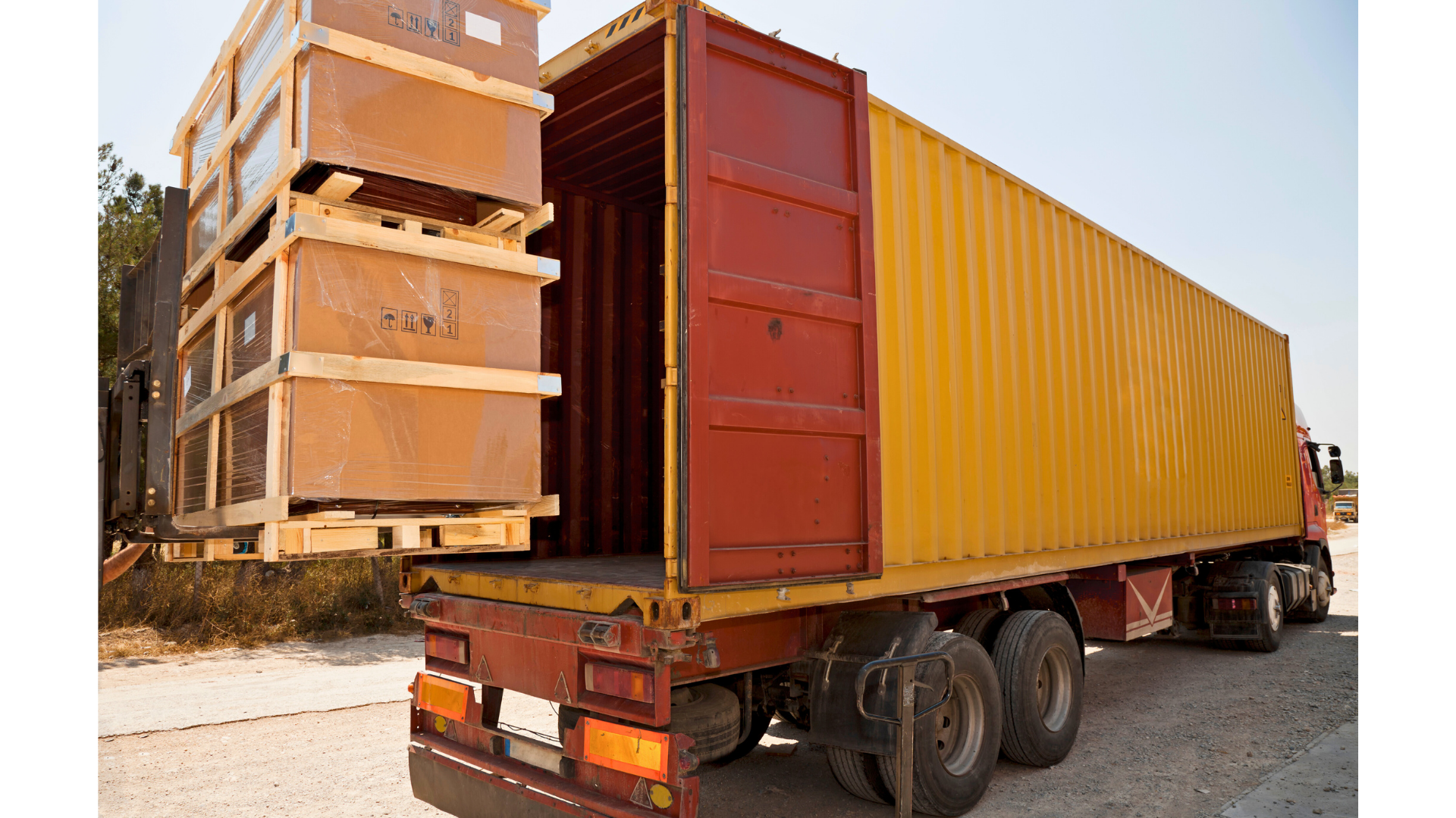 A yellow shipping container truck with red accents is parked outdoors. The container is open, and several large wooden crates are being loaded onto it, with some crates wrapped in plastic.