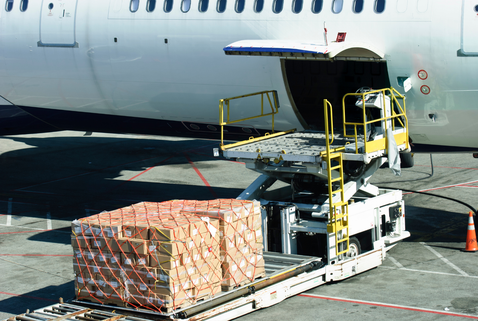 Cargo loading equipment beside an airplane, with a black platform with yellow safety rails and a table with orange netted pallets of cargo.