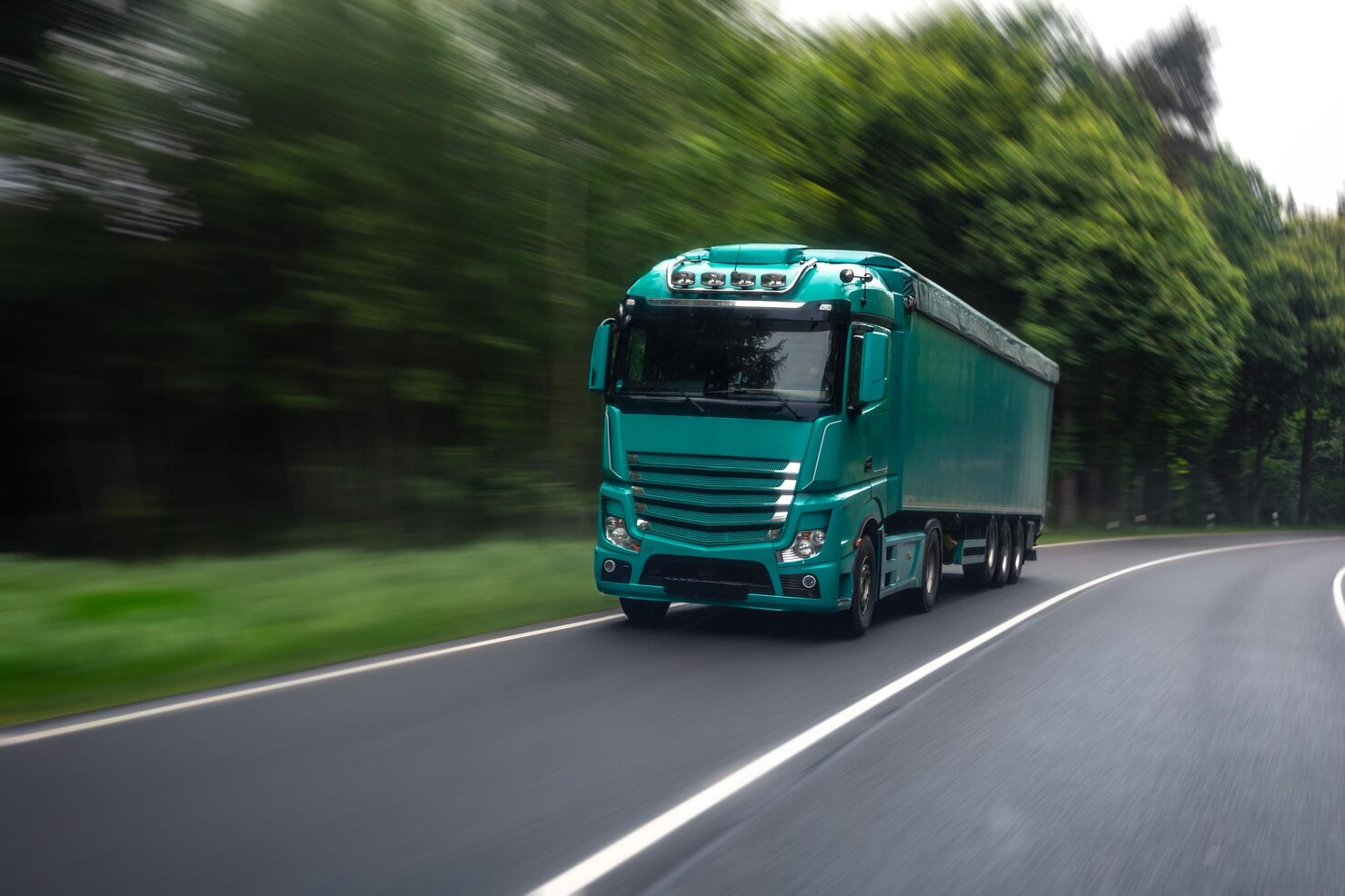 A teal semi-truck driving on a curved road surrounded by green trees.
