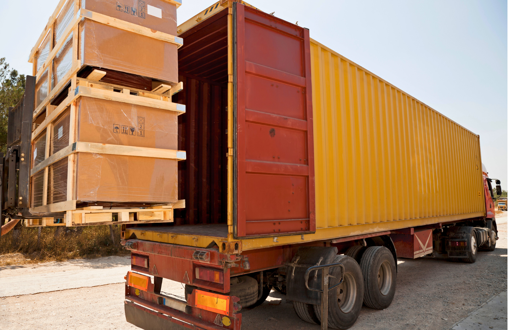 A large yellow transport truck with an open back, carrying wooden crates and boxes loaded with large items, some wrapped in plastic, in an outdoor setting.