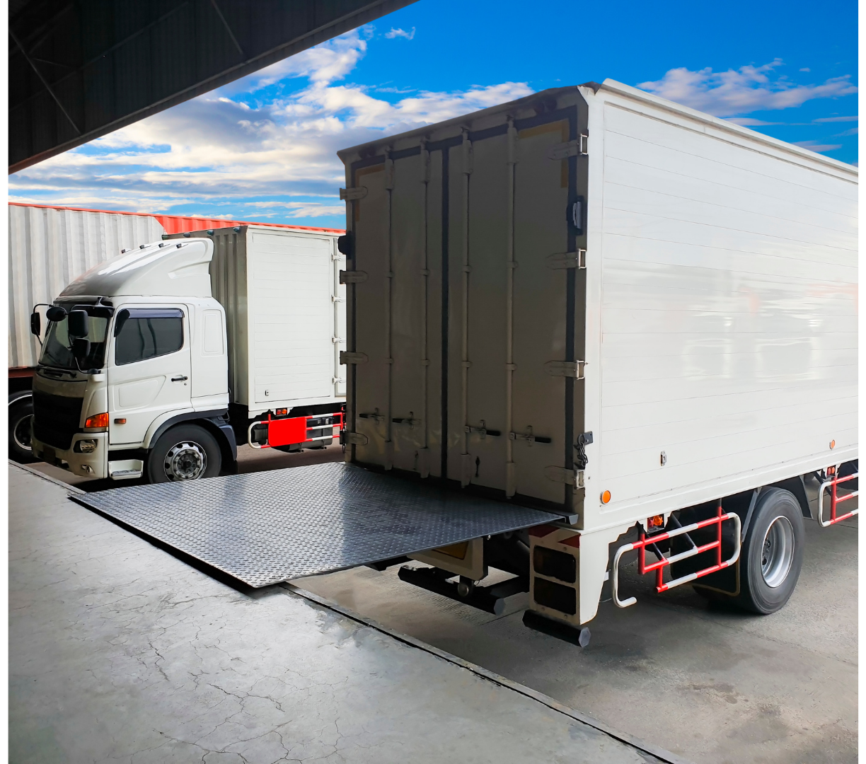 A white delivery truck with a loaded cargo area is parked inside a warehouse. The truck has a fold-down ramp at the rear, leading to the ground outside, with a blue sky and scattered clouds overhead.