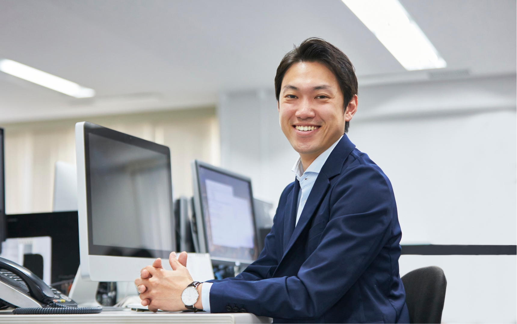 A young man in business attire sitting at a desk in an office. He is smiling and looking at the camera with hands clasped. On the desk, there is a computer monitor, a telephone, and some office supplies. The background shows additional computer monitors and office lighting.