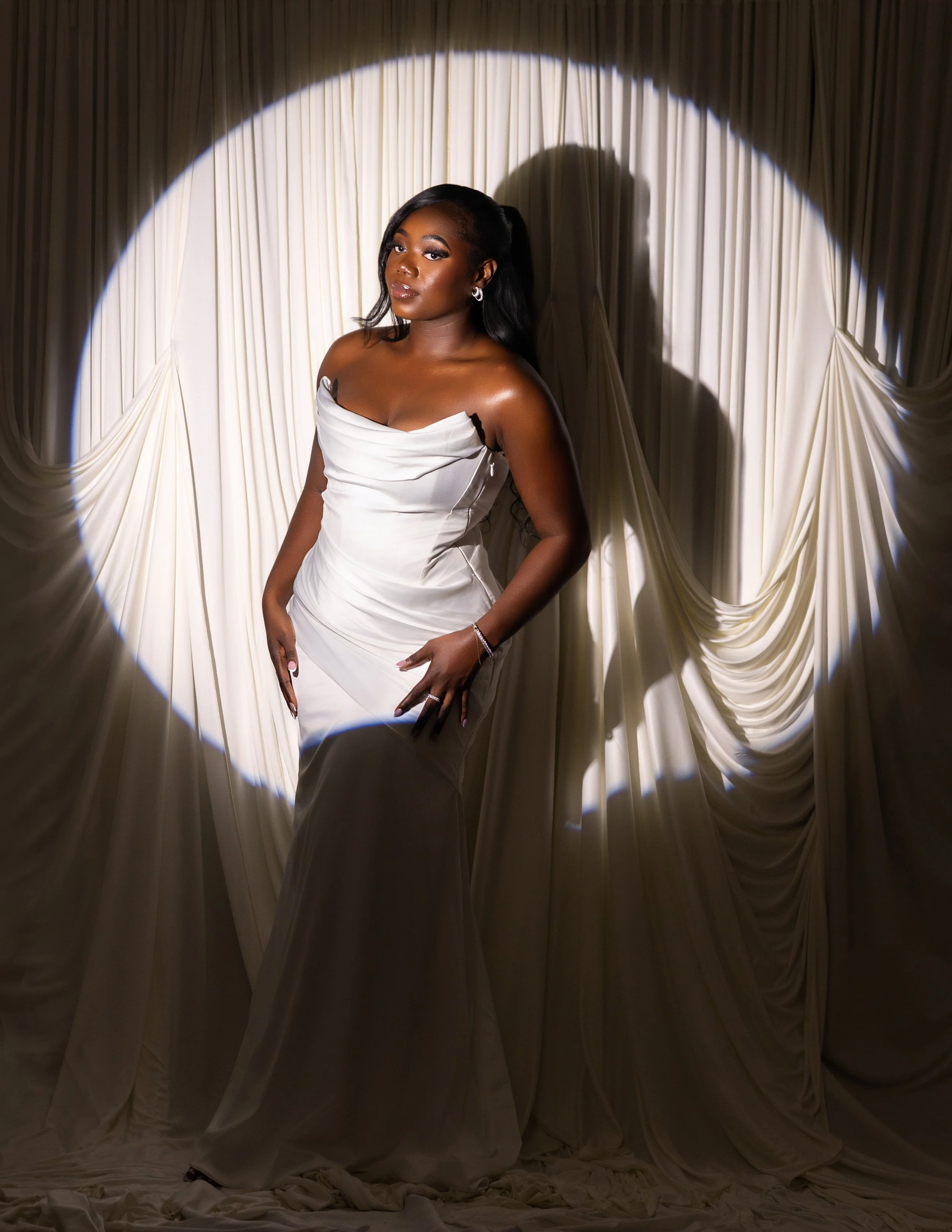 A woman in a white dress standing in front of a cream-colored draped curtain, illuminated by a circular spotlight.