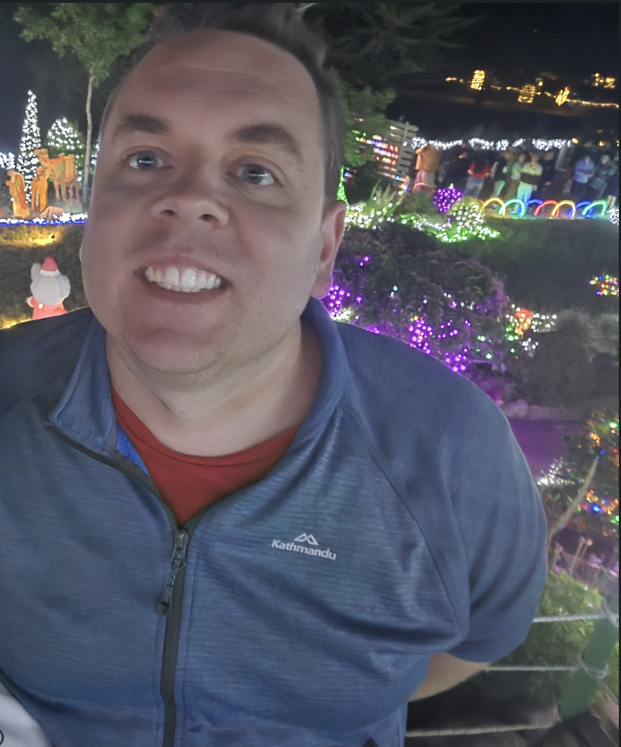 A man smiling at a nighttime outdoor holiday light display with decorated trees, illuminated decorations, and a crowd of people in the background.