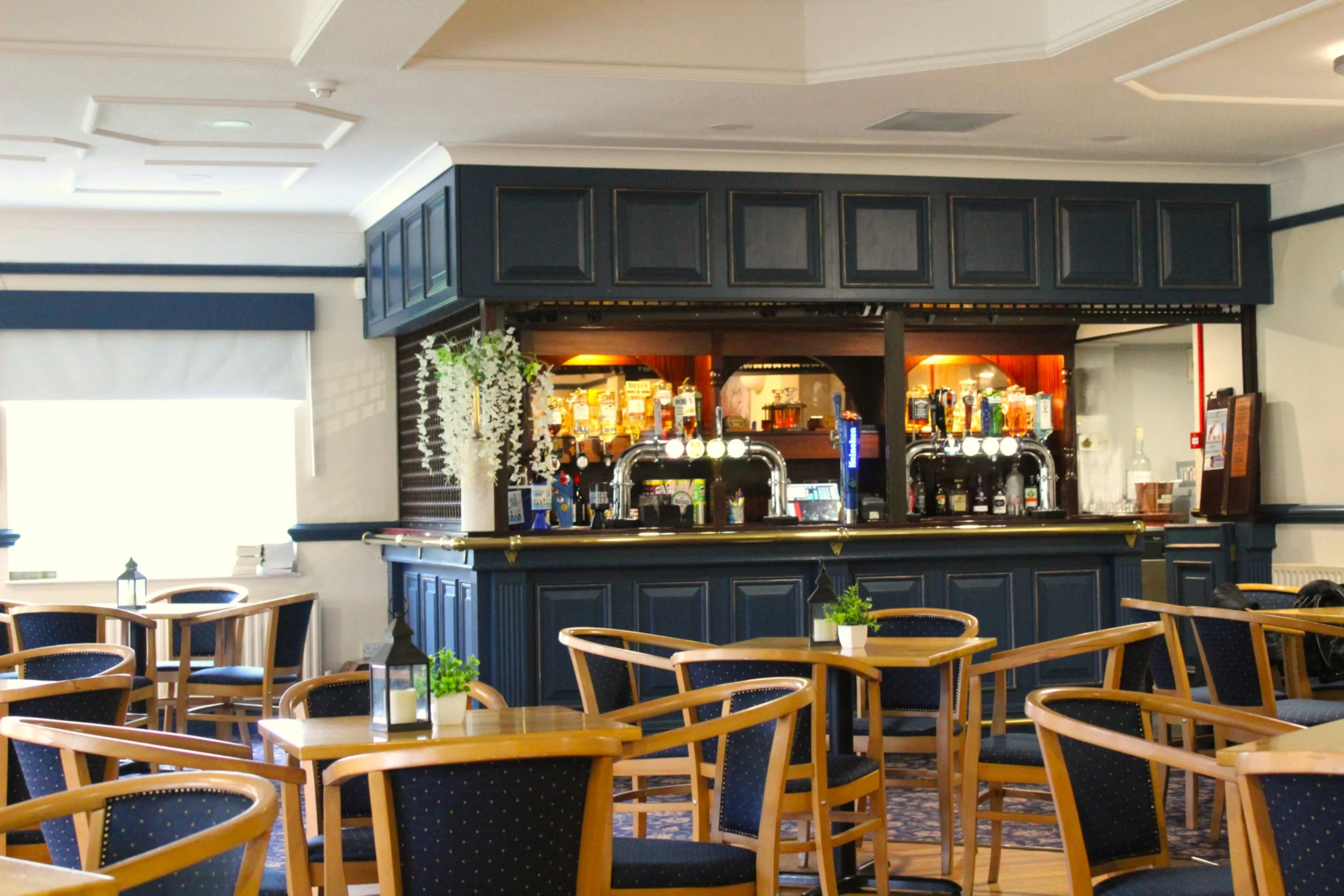 Interior view of a restaurant or cafe with multiple wooden tables and chairs with blue upholstery. A bar area with liquor bottles and beer taps is visible in the background. The room has white walls, decorative molding, and natural light coming from windows.
