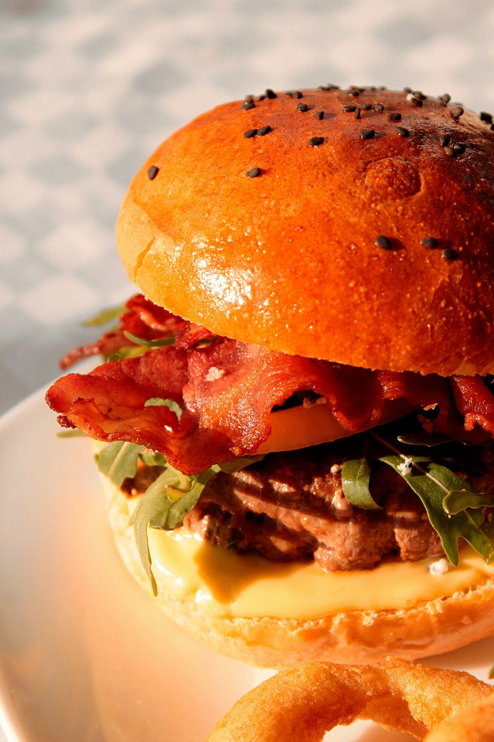 Close-up of a gourmet burger with bacon, arugula, tomato, cheese, and a toasted bun topped with black sesame seeds, served with a side of onion rings.