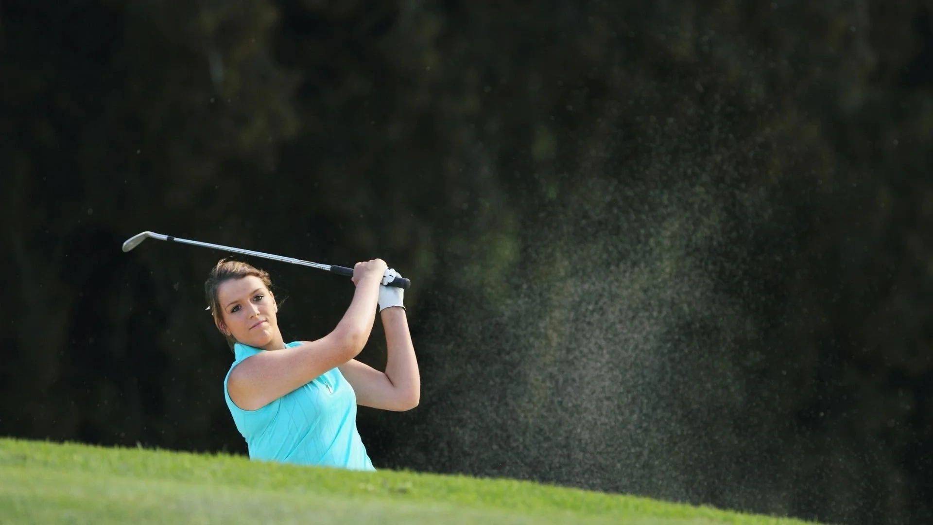 Female golfer in a blue sleeveless top swinging her golf club on a golf course.