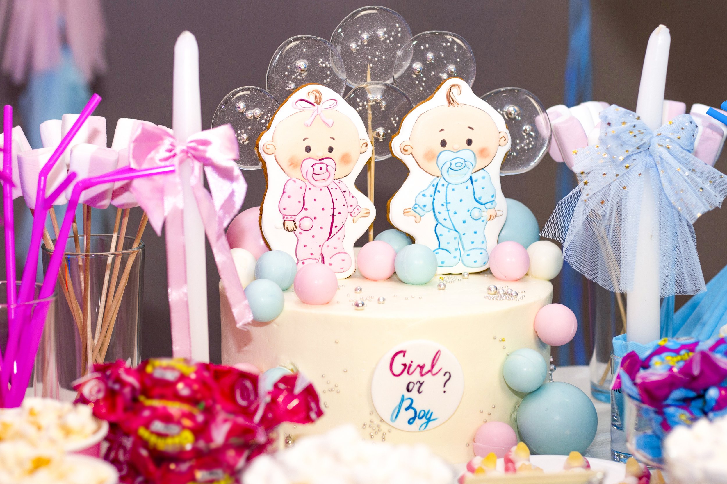 Decorated cake with pink and blue baby-themed cookies, surrounded by pink, blue, and purple candies and party decorations for a gender reveal party.
