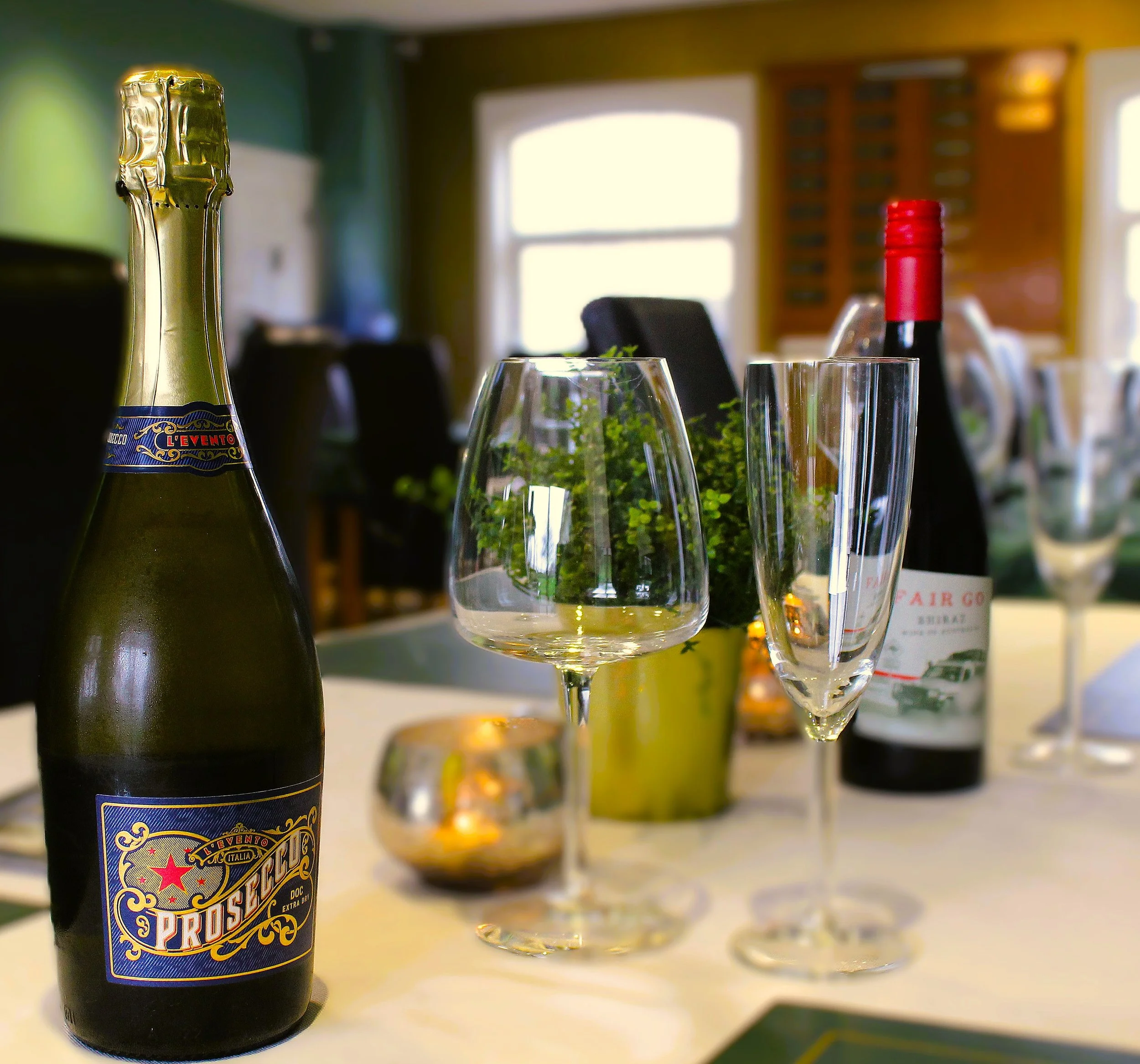 A bottle of sparkling wine, a glass of white wine, and a glass of red wine on a table in a bright room with large windows and wooden cabinets in the background.