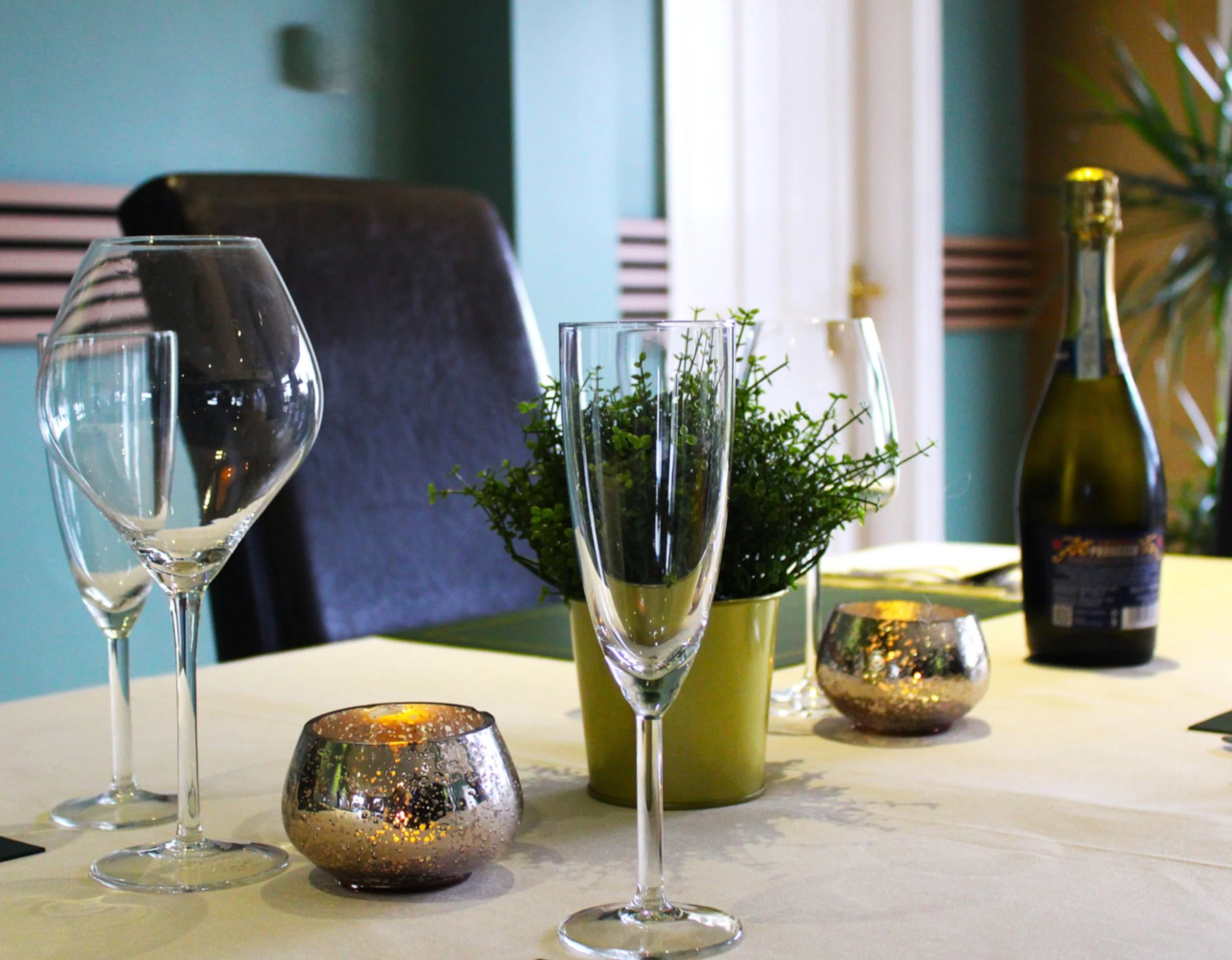 Elegant table setting with empty wine glasses, decorative candles, potted plants, and a bottle of beverage in a well-lit room with teal walls and curtains.