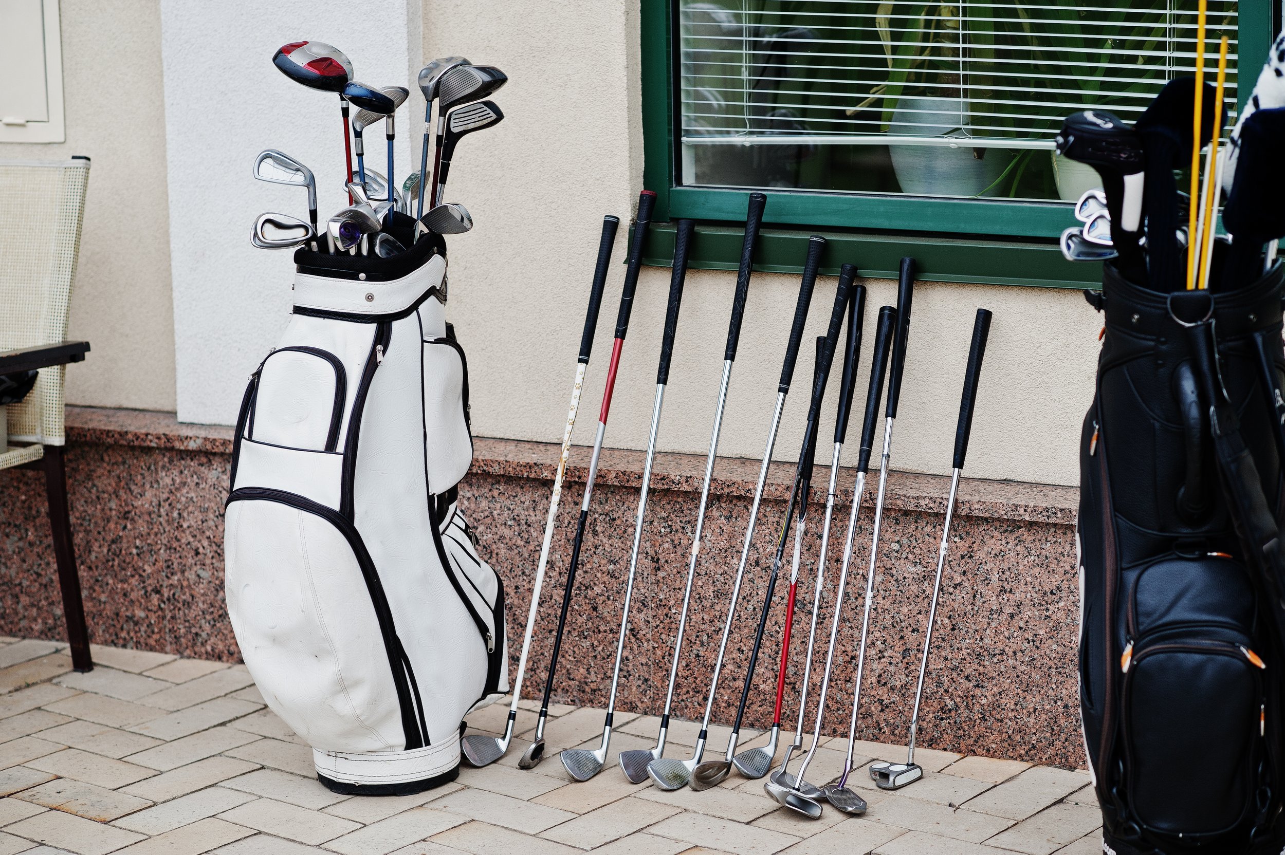 Golf clubs in golf bags and extra clubs lined up outside near a wall and window.