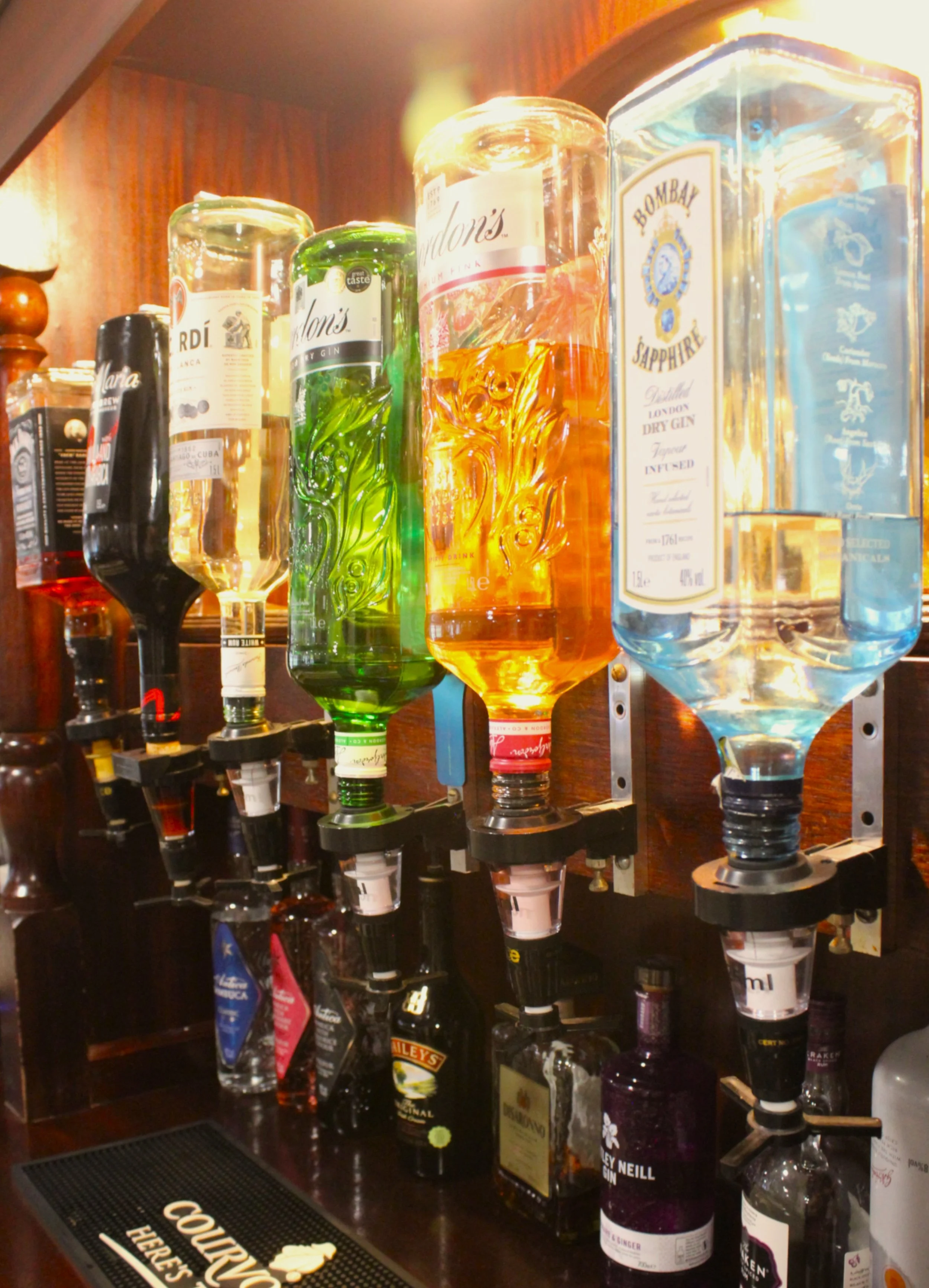 Various bottles of gin with pouring spouts on a bar shelf, including Bombay Sapphire, No. 209 Dry Gin, and Hendrick's, behind a black bar mat.