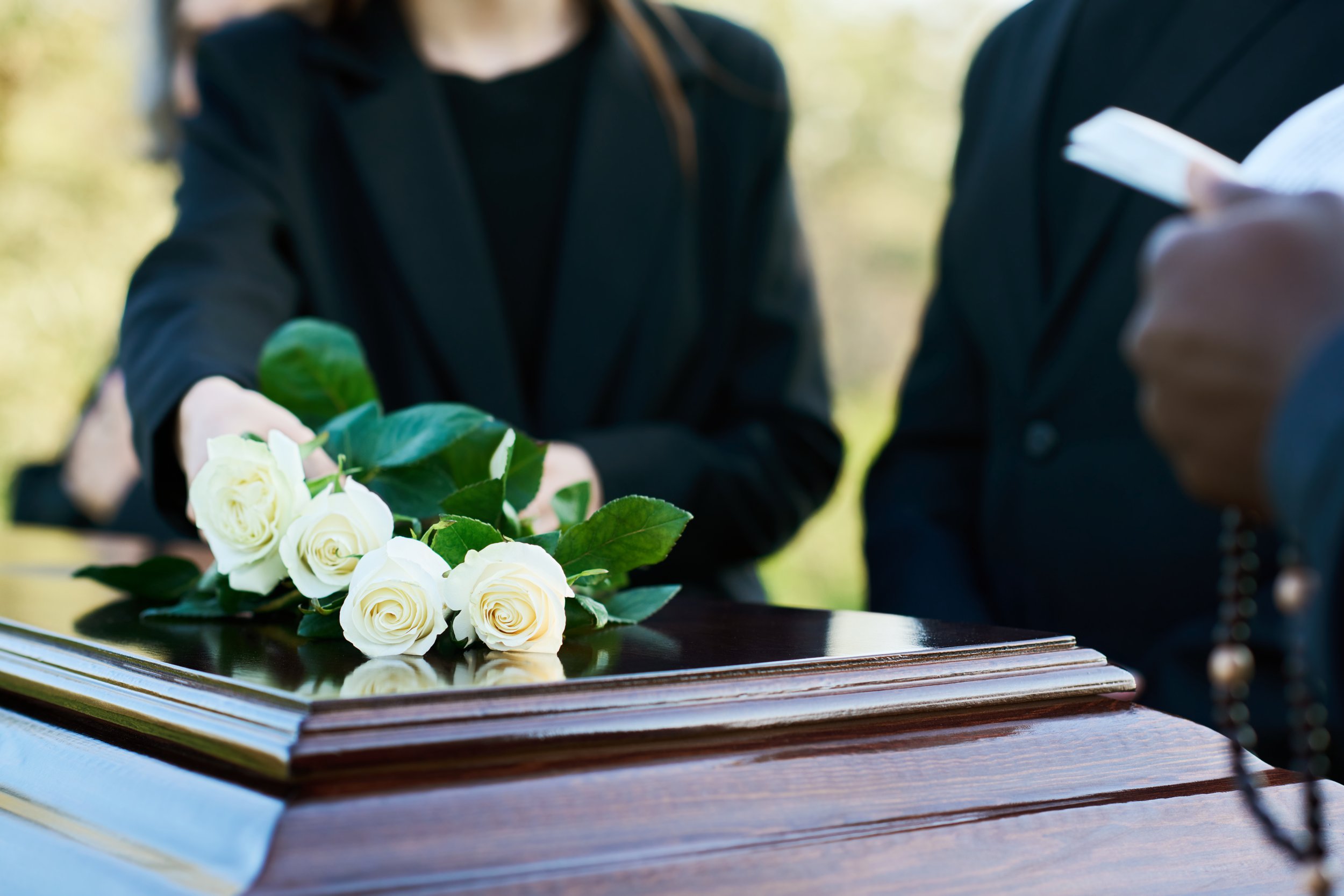 A bouquet of white roses and green leaves placed on an open fashion magazine on a wooden table. Blurred people in dark clothing are in the background, with one person holding a rolled-up magazine or paper.