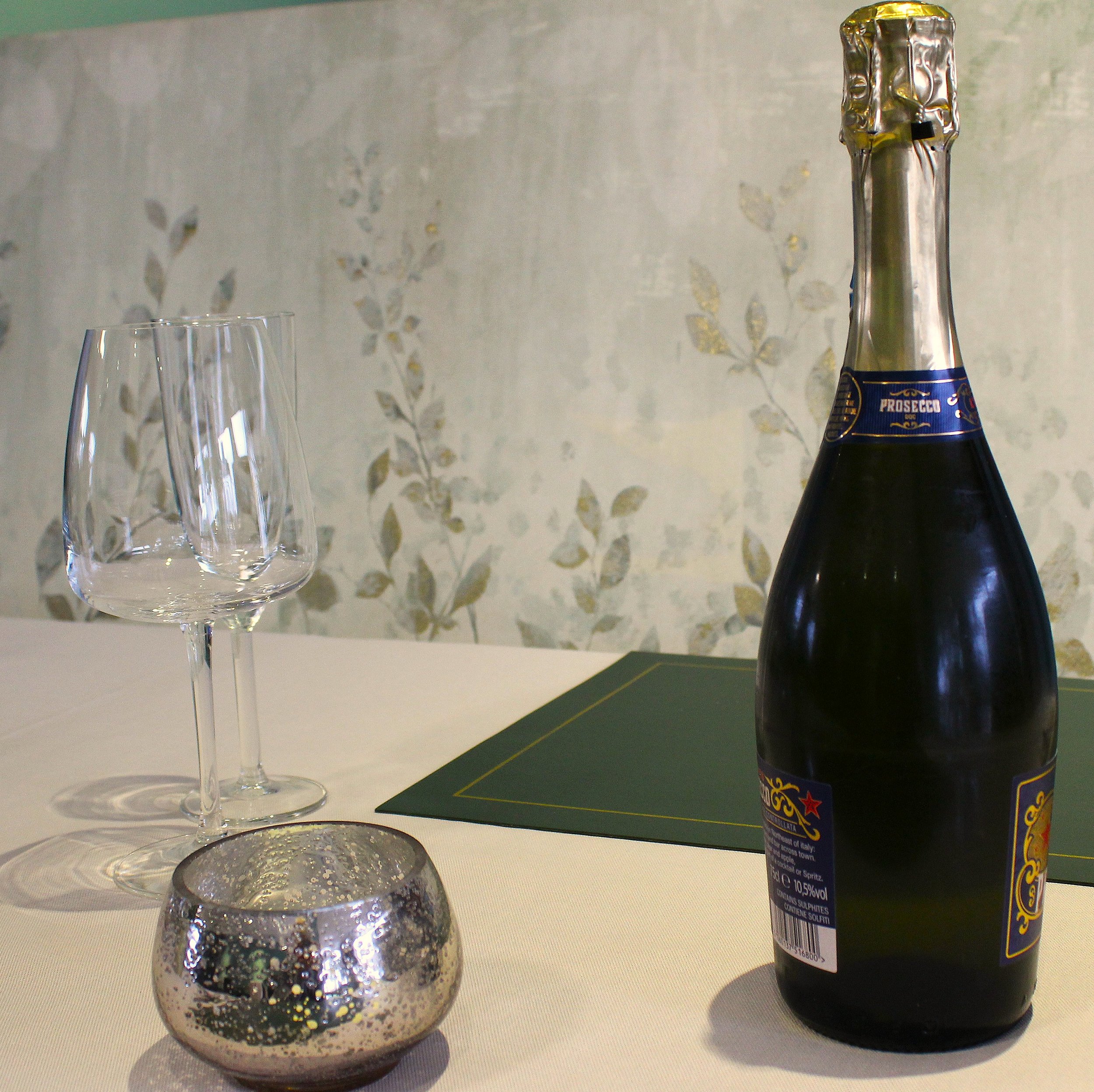A clear glass filled with glittery silver and black confetti next to a dark bottle of sparkling wine or champagne, on a white surface against a plain wall background.