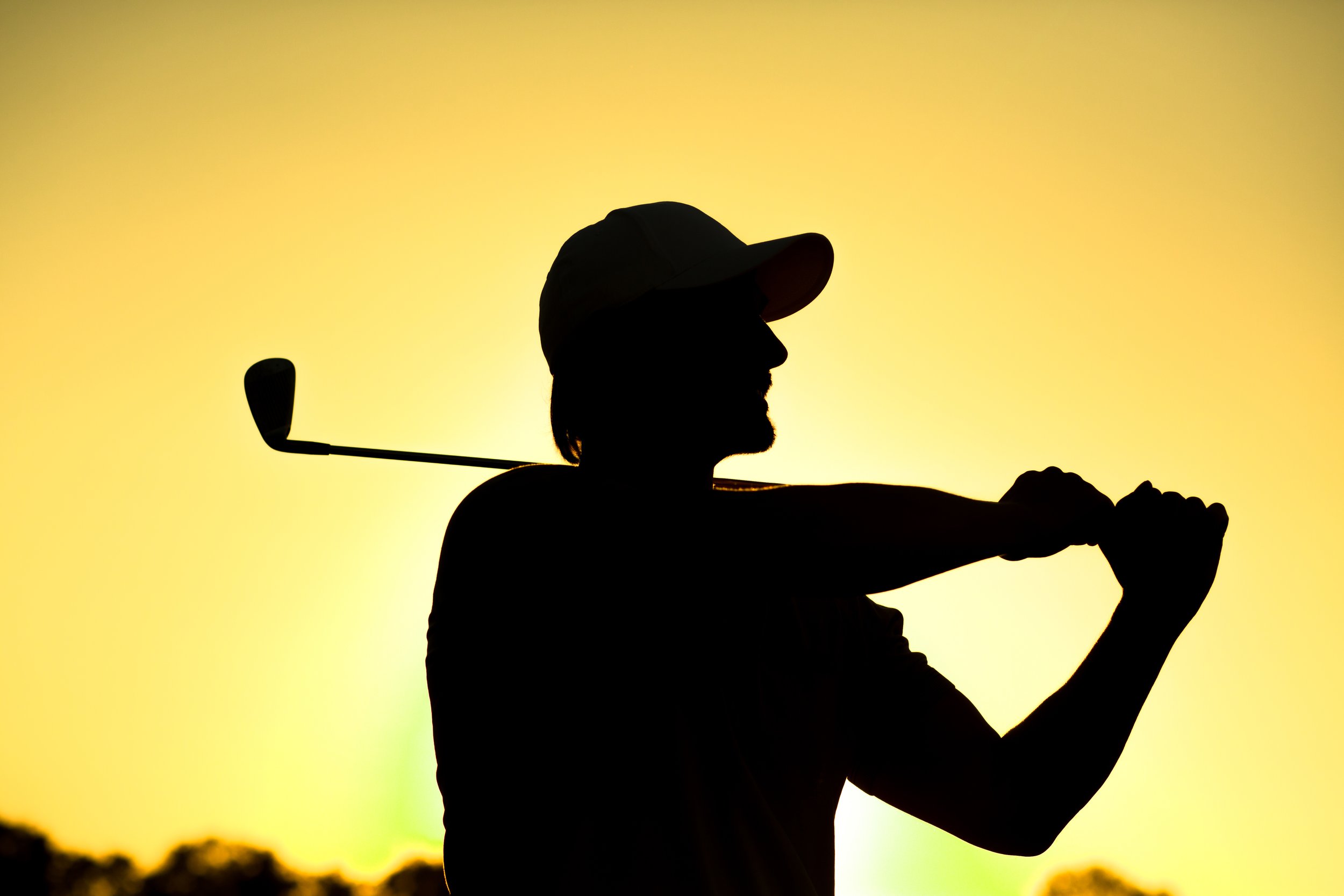 Silhouette of a person wearing a cap, holding a golf club, with a sunset or sunrise in the background.