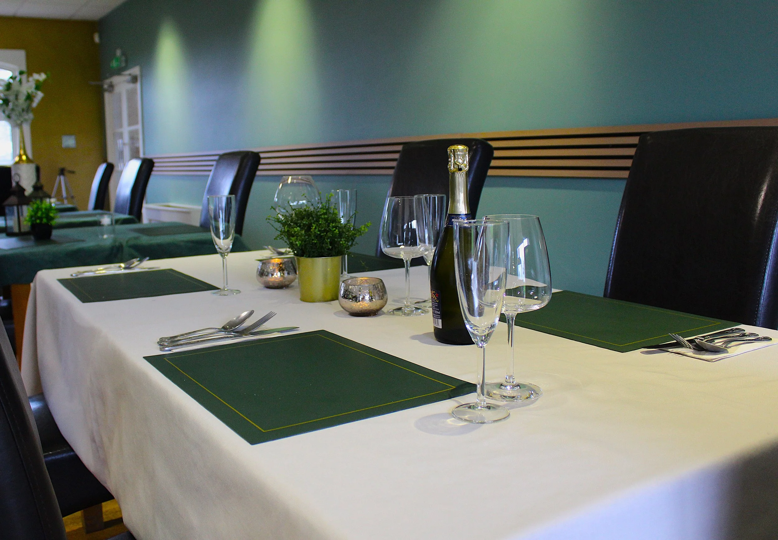 A dining table with black chairs, set with champagne bottles, glasses, plates, silverware, and small potted plants, in a room with a teal-colored wall and green accent decor.