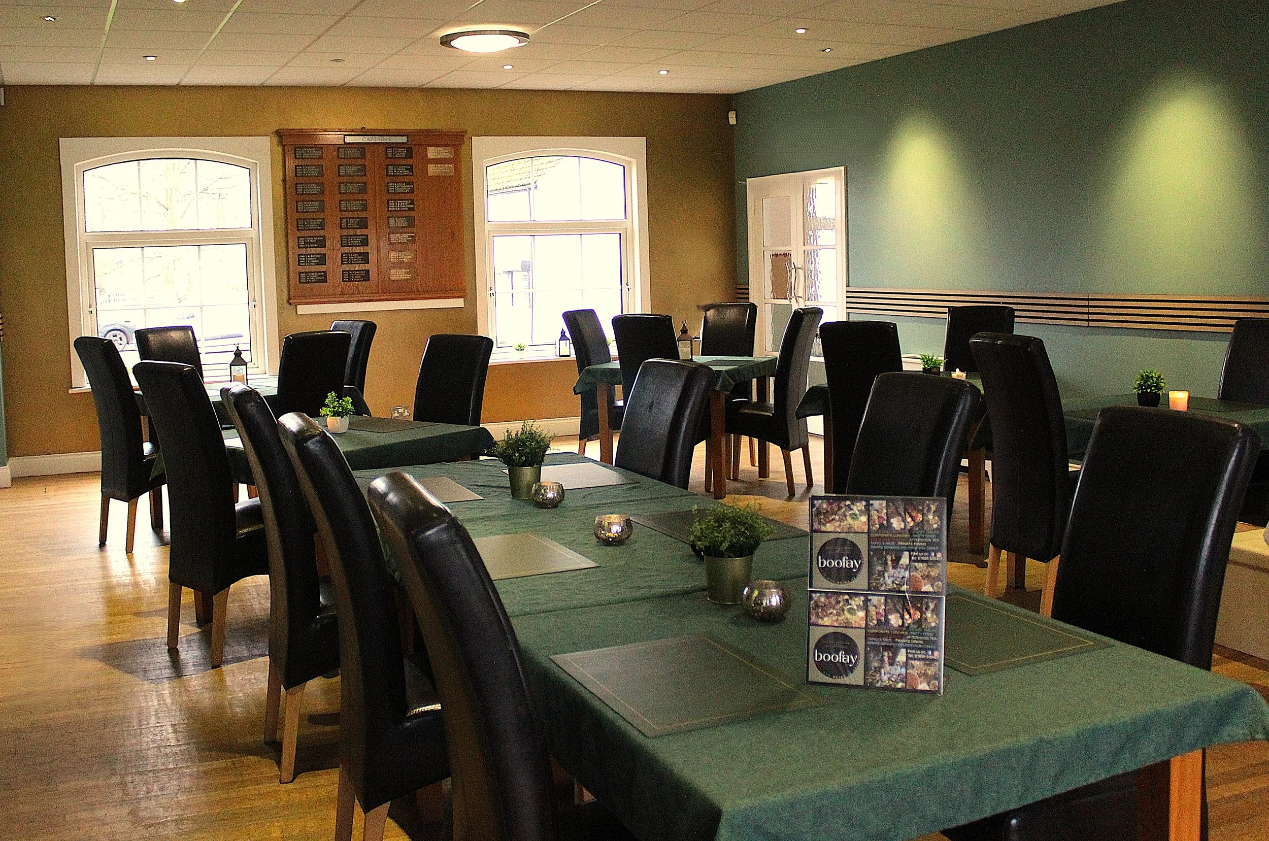 Interior of a restaurant or café with dark wood tables, black chairs, and small potted plants. Bright windows with natural light, and a board on the wall with several black plaques. A table at the foreground has small candleholders and a menu or brochure promoting a brand called 'boofay'.