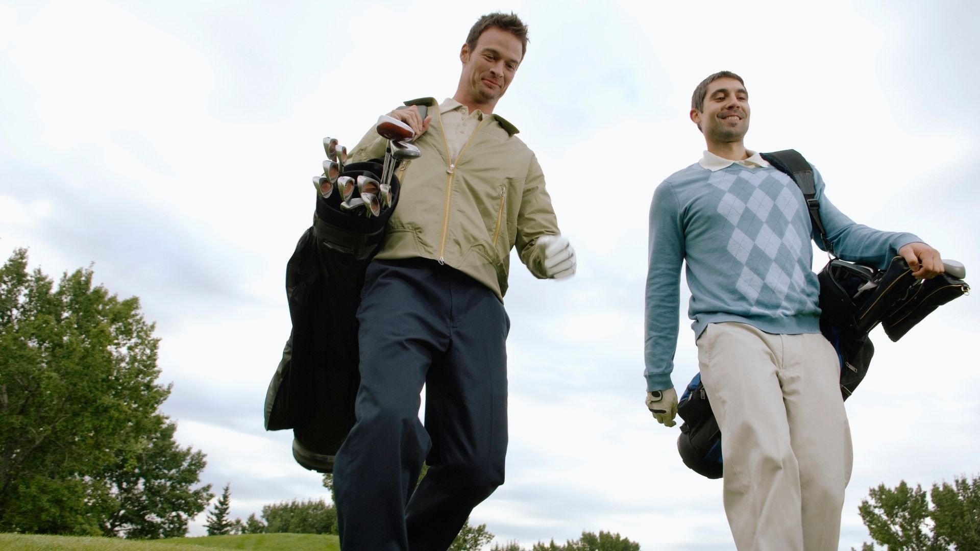 Two men walking on a golf course carrying golf clubs in their bags, smiling and enjoying a day of golf.