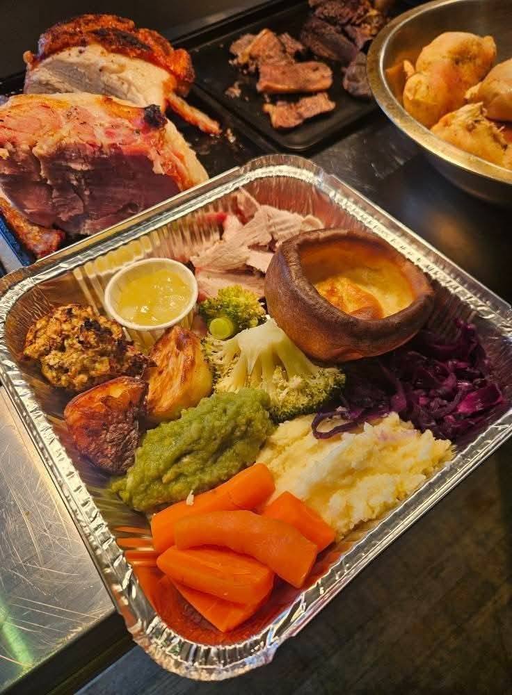 A foil tray with assorted cooked vegetables including carrots, mashed potatoes, broccoli, roasted sweet potato, red cabbage, and a small bowl of gravy or sauce, along with slices of ham, roast chicken, and beef in the background.