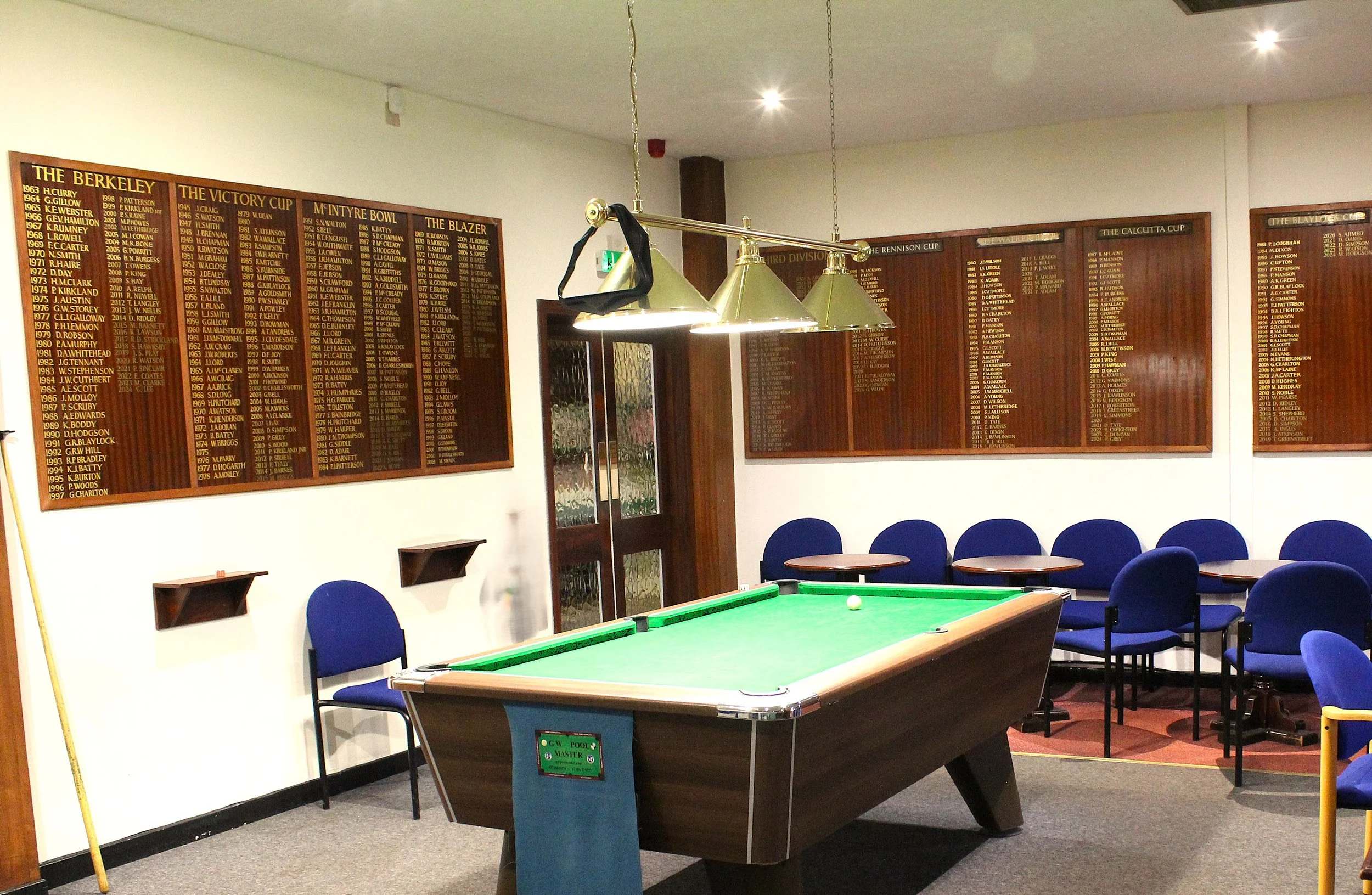 An indoor recreation room featuring a pool table, blue chairs, and large wooden plaques on the wall listing sports achievements.