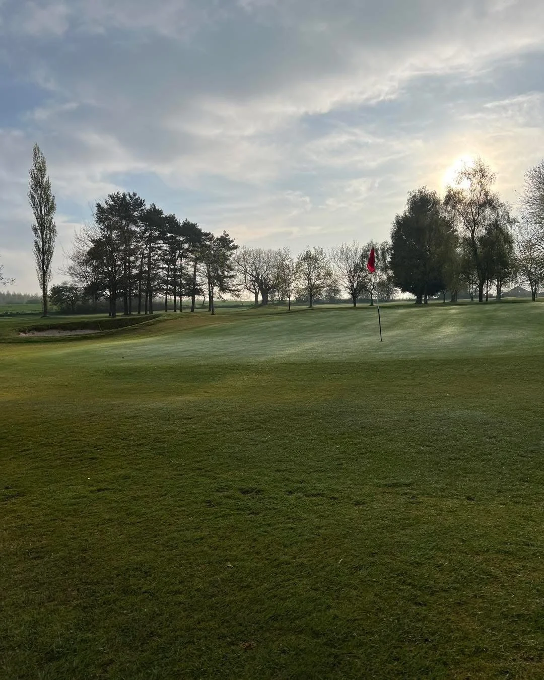 A golf course with a putting green and flag, surrounded by trees under a partly cloudy sky at sunset.