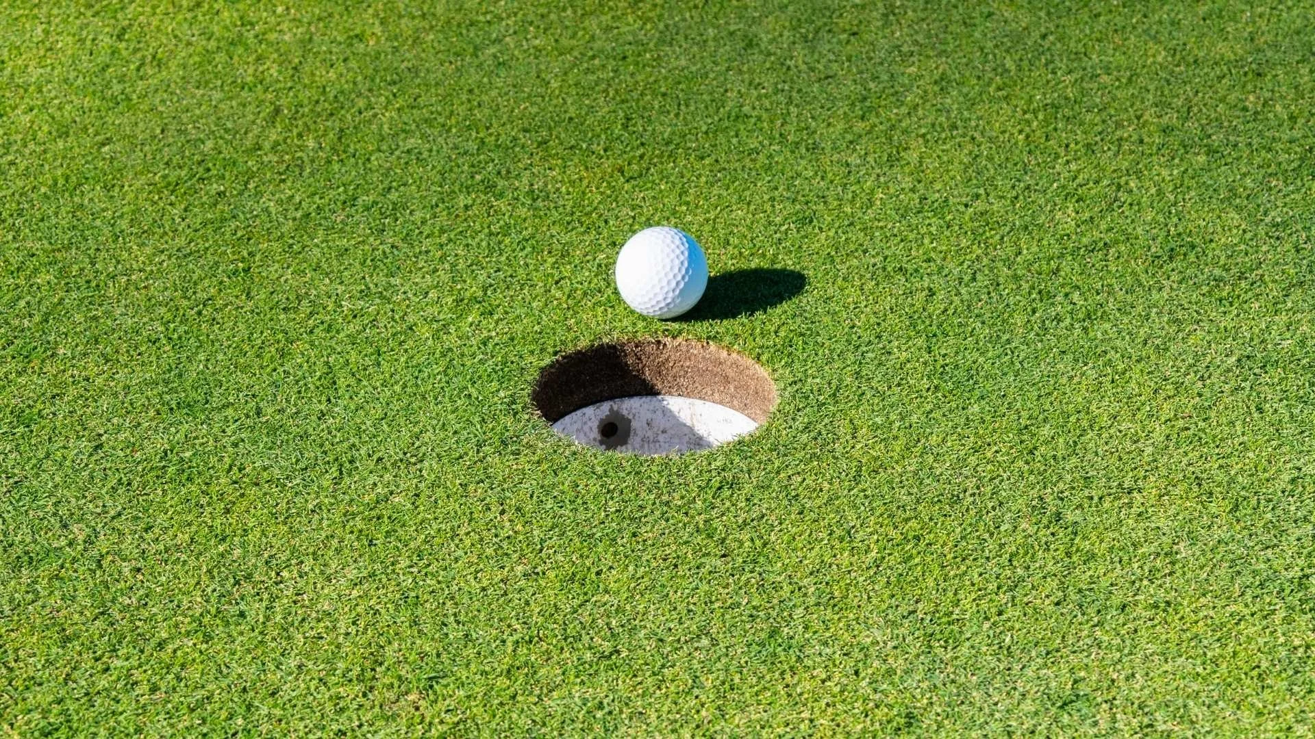 A golf ball resting near the edge of a golf hole on a green, with bright sunlight casting a small shadow of the ball onto the grass.