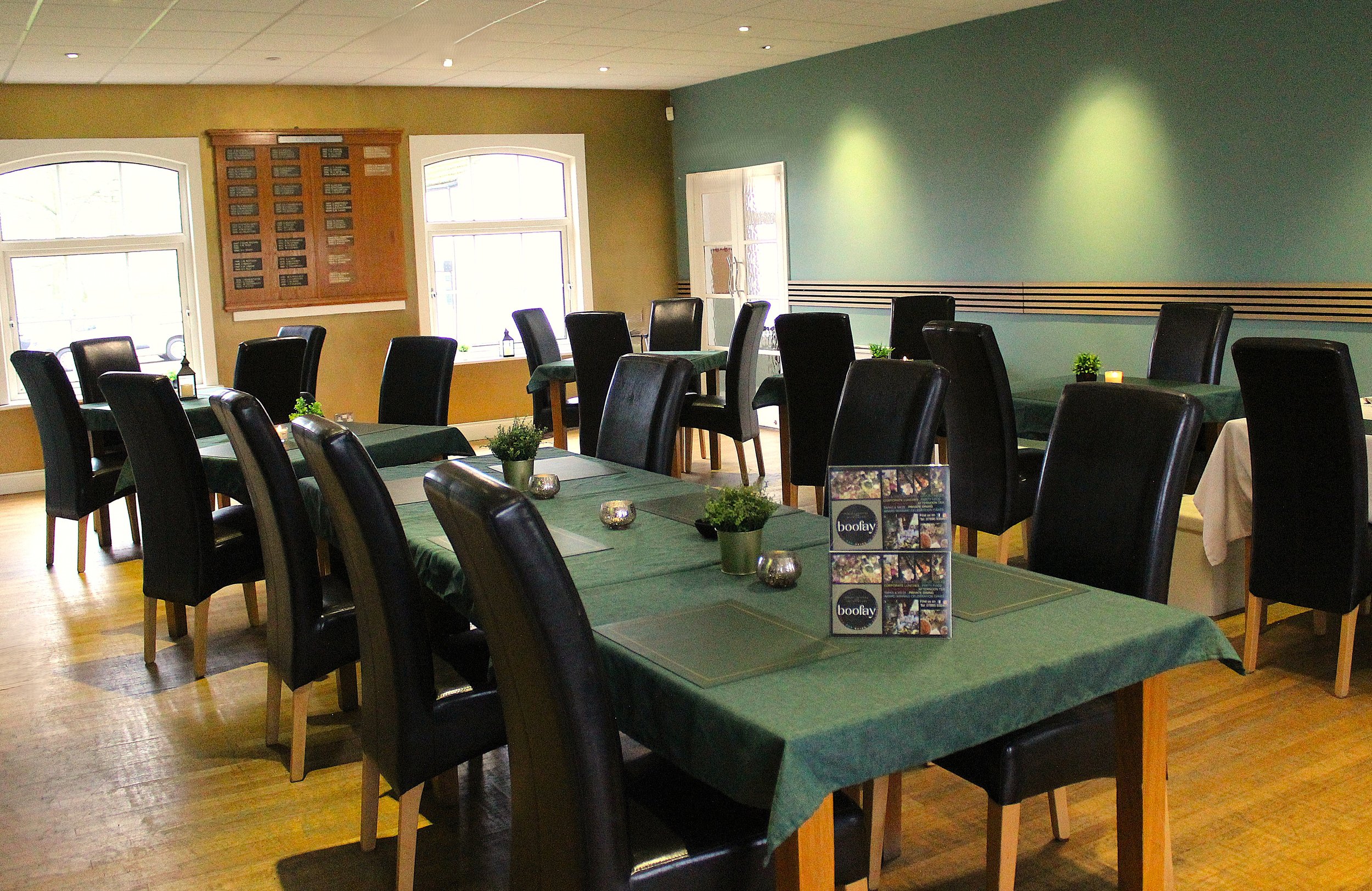 Interior of a restaurant or cafe with green tablecloth-covered tables, black chairs, small plants, candles, and candleholders. There are large windows, wooden wall panels, and a multi-pane window with a door in the background, all with natural light.