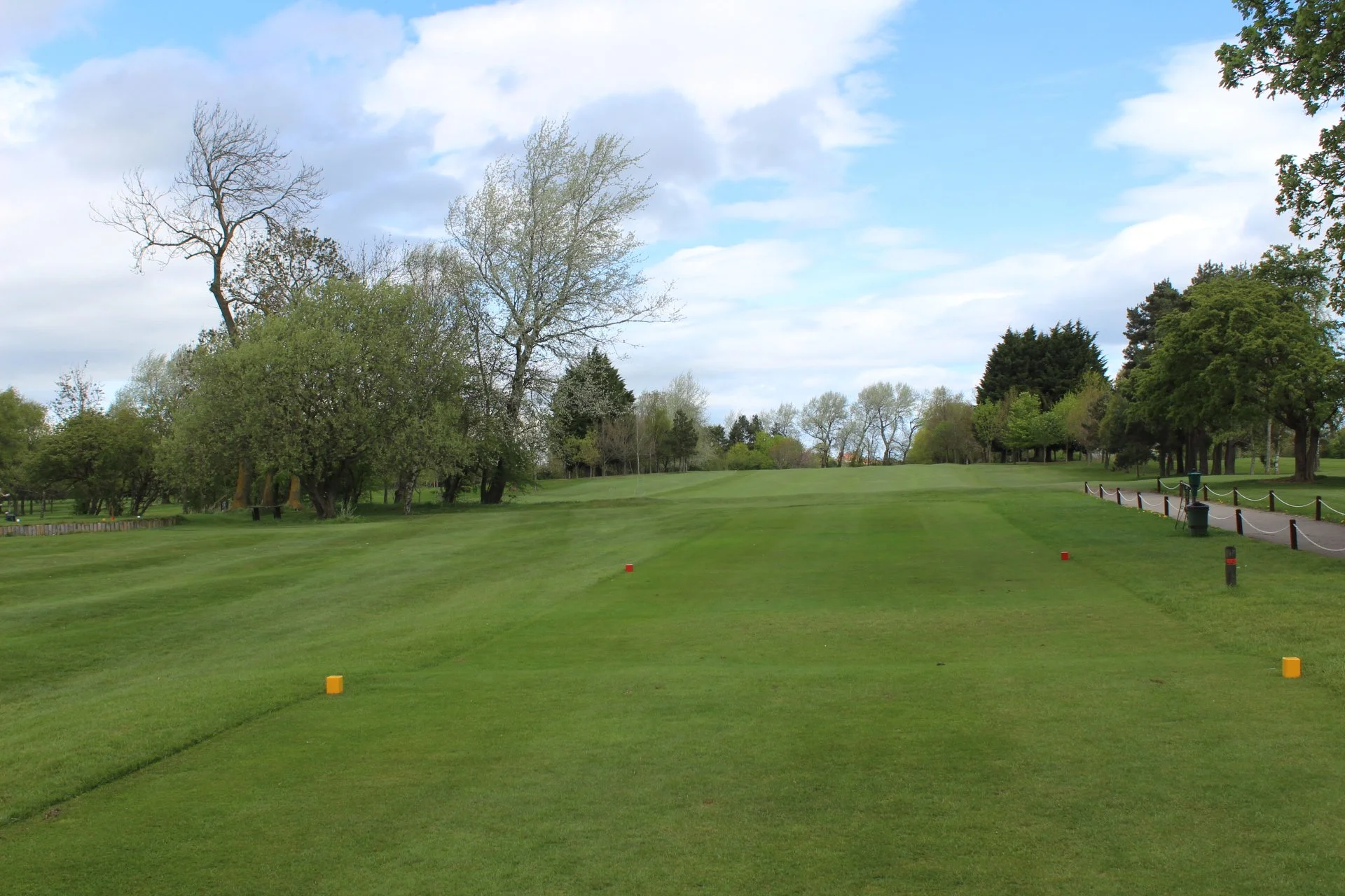 A scenic view of a golf course with lush green grass, scattered trees, a partly cloudy sky, and a paved pathway with black posts and chain links.