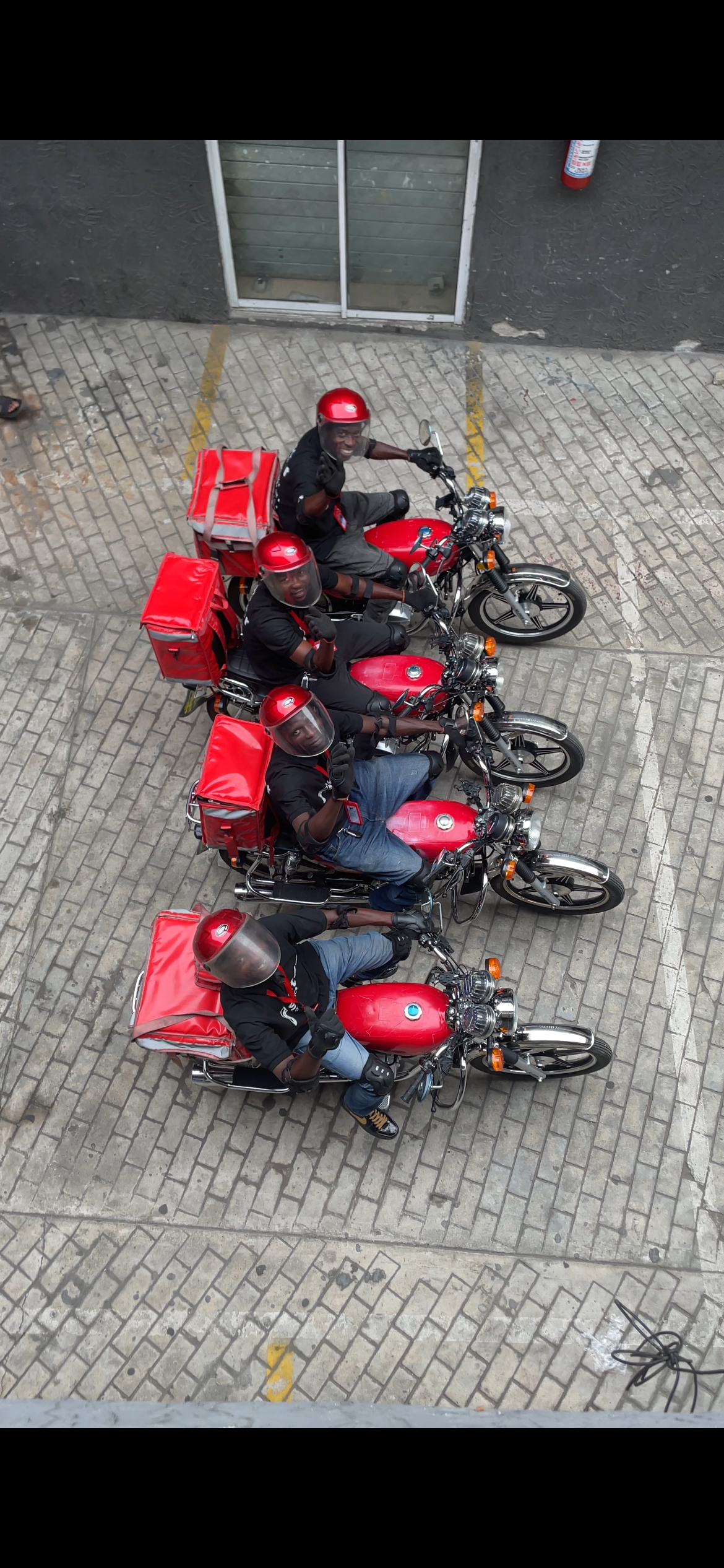 Four delivery drivers sitting on red motorcycles, wearing red helmets and black uniforms, parked on a brick-paved street.