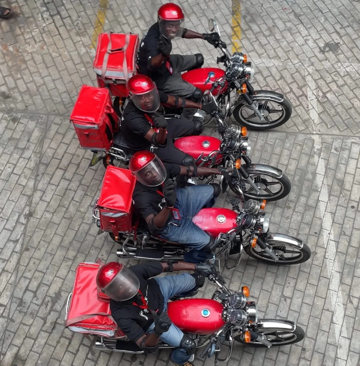 Five motorcyclists wearing red helmets and black shirts, sitting on red motorcycles with red delivery bags attached to the back, viewed from above on a cobblestone street.